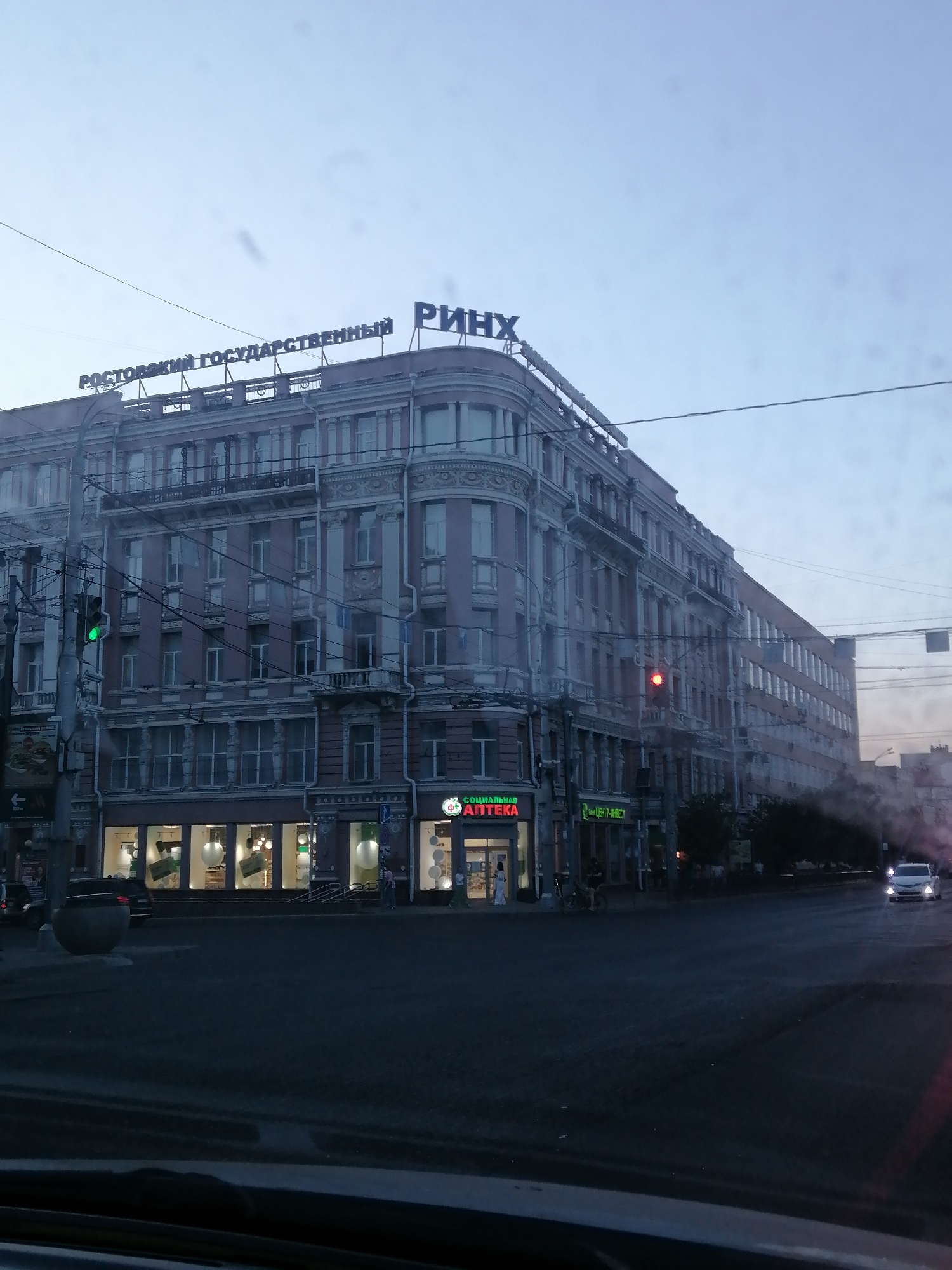 The image depicts a street scene at dusk in Rostov-on-Don, Russia. Dominating the frame is a large, ornate pink building with multiple stories, featuring numerous windows and decorative architectural elements. On top of the building, large blue letters spell out "РОСТОВСКИЙ ГОСУДАРСТВЕННЫЙ РИНХ," indicating that this is the Rostov State University of Economics (RSUE), commonly known as RINKh.
Below the main building, a row of storefronts lines the ground floor, with illuminated windows suggesting businesses are open. A sign for "СОЦИАЛЬНАЯ АПТЕКА" (Social Pharmacy) is visible on the left. Traffic lights are present, with one showing a green light and another further down the street showing red.
The street in front of the building appears to be an intersection or a wide road. A few cars are visible, including a white car driving away in the distance. The overall atmosphere is dim, consistent with twilight. The image appears to be taken from inside a vehicle, looking out through a window, as evidenced by the slight reflections and the framing of the foreground. The sky is a pale, overcast blue.