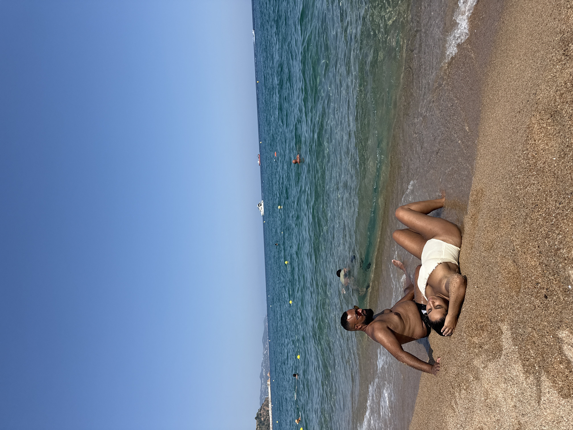 This image depicts a sunny day at a beach in Blanes, Spain. In the foreground, a couple is relaxing on the sandy shore. A man with dark hair and a beard, wearing sunglasses and black swim trunks, is seated with his legs extended towards the water. Next to him, a woman is lying down on her back, wearing a white swimsuit.
The ocean stretches out before them, with clear, turquoise water. Several other people are visible in the water, some swimming and others relaxing. Yellow buoys are scattered across the water, marking a designated swimming area. In the distance, a white yacht and a few other boats are sailing on the horizon. The coastline is visible in the background, with hilly terrain and buildings dotting the landscape. The sky is a clear, bright blue, indicating good weather. The overall scene conveys a peaceful and leisurely atmosphere on a summer day.