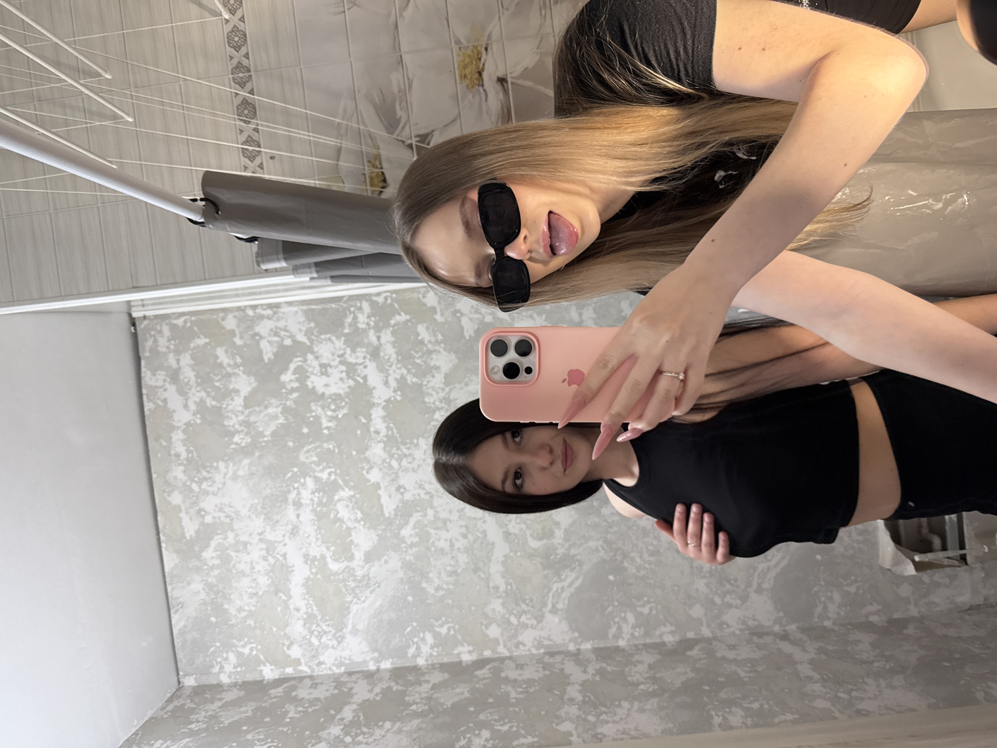 Two young women are posing for a selfie in what appears to be a bathroom. The woman in the foreground has blonde hair and is wearing sunglasses, sticking out her tongue. The second woman, presumably taking the photo, has dark hair and is wearing a black top. The environment includes a bathroom with patterned wallpaper, a shower curtain, and a drying rack. The location is Chaikovsky, Russia.