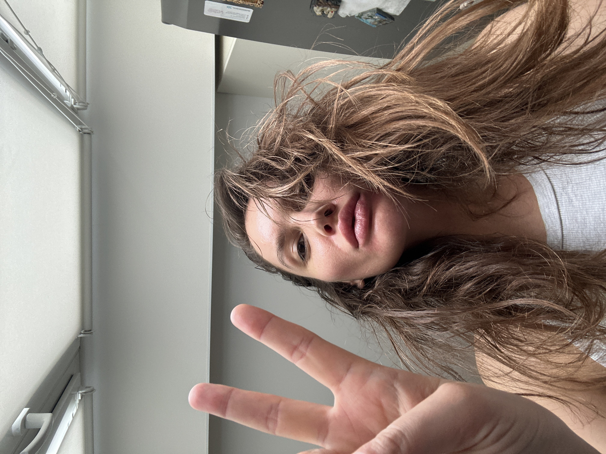 This image is a selfie of a young woman taken indoors in Moscow, Russia. The woman has long, light brown, wavy hair that appears voluminous and somewhat disheveled, falling around her face and shoulders. Her head is tilted to the right, and she looks directly into the camera with a neutral to slightly pensive expression, her full lips slightly parted. She is wearing a light-colored, possibly grey, top.
In the immediate foreground, her right hand is raised, making a peace sign (V-sign) with her index and middle fingers extended. The background primarily consists of a plain white wall, suggesting an indoor setting. On the left side of the frame, a light-colored, possibly metal or plastic, structure resembling part of a window frame or a small rack is visible. In the upper right corner, above the woman's head, there is a glimpse of a darker surface, possibly a shelf or cabinet, on which a few small, indistinct objects rest. One of these objects appears to have faint, unreadable text or a logo. The lighting is bright and diffuse, indicating it is likely daytime. The overall image is rotated 90 degrees clockwise, presenting an angled perspective.