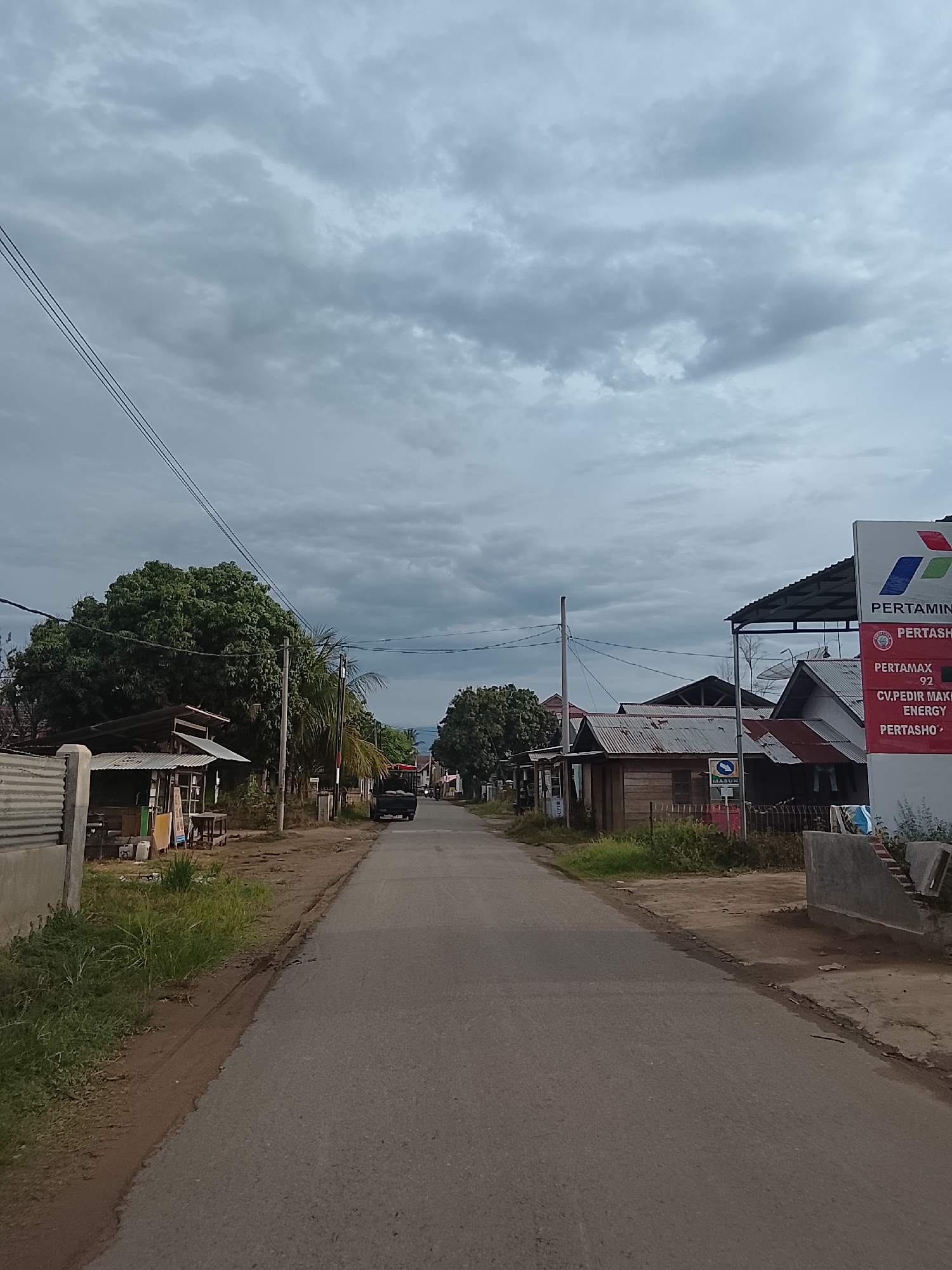 The image shows a street scene in Geunteng, Indonesia, under a cloudy sky. The street is paved and appears to be a rural or suburban road. On the left side of the road, there are several small, simple buildings with corrugated metal roofs, some with fences and overgrown vegetation. A large, leafy tree is prominent on the left. On the right side of the road, a tall sign for Pertamina, a state-owned oil and gas company, is visible, along with more buildings with corrugated roofs. A dark-colored truck is driving away from the viewer down the street. The environment appears to be somewhat undeveloped, with a mix of structures and natural elements.