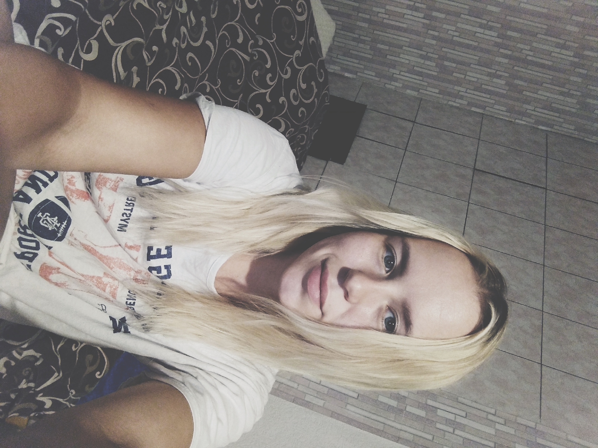 A young blonde woman with blue eyes is taking a selfie in a room. She is wearing a white t-shirt with a graphic design. She is sitting on a bed with a dark patterned comforter. The wall behind her is tiled with a brick pattern.