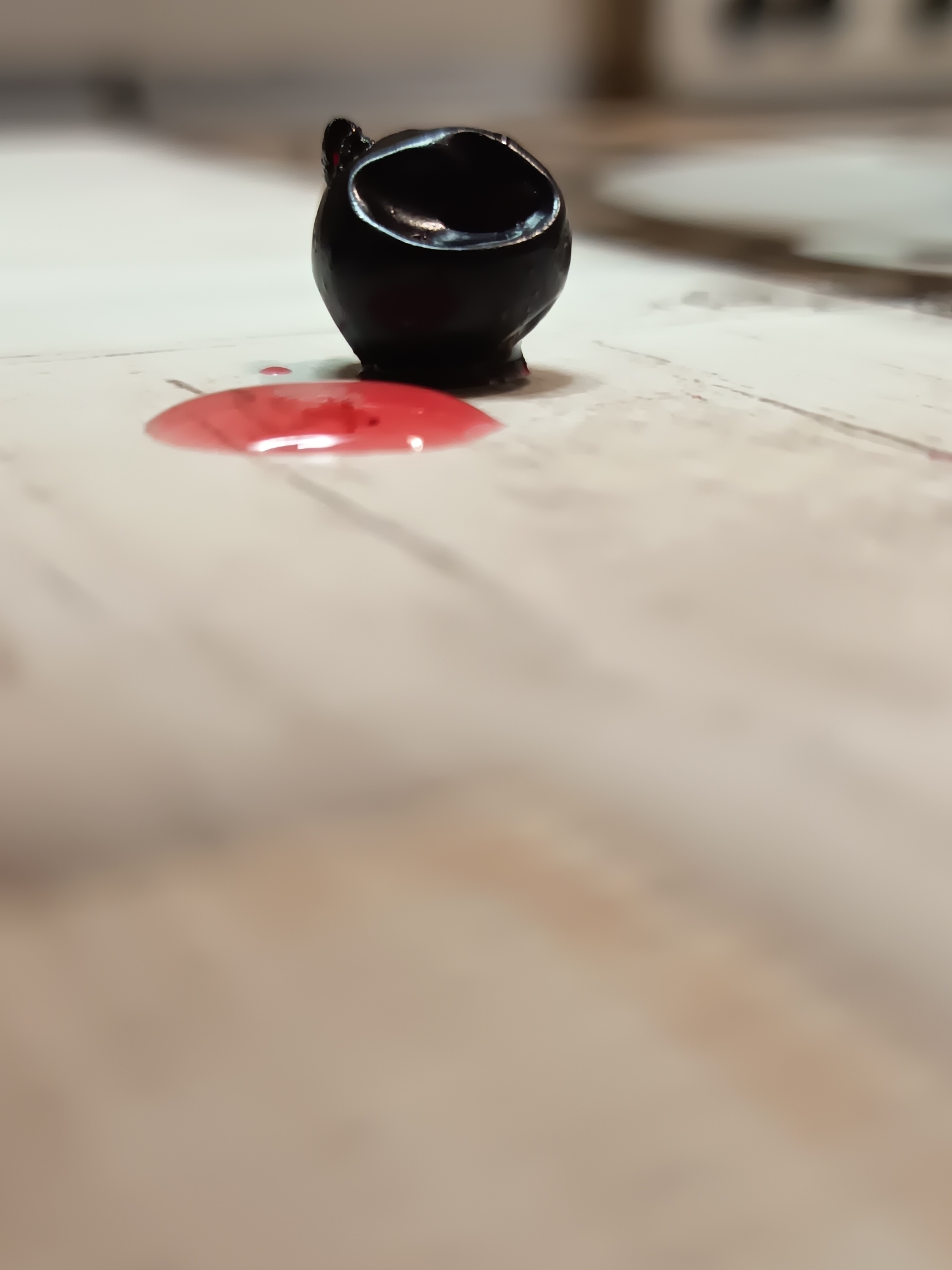A macro shot of a black, ceramic ink pot tilted on its side, with a small puddle of red ink spread on a light-colored, textured surface that appears to be a wooden table. The background is blurred, with hints of off-white and light brown, suggesting an indoor setting. The image focuses on the spilled ink and the ink pot, creating a sense of drama or narrative. No specific location in Izmail, Ukraine, is identifiable from the visual information.
