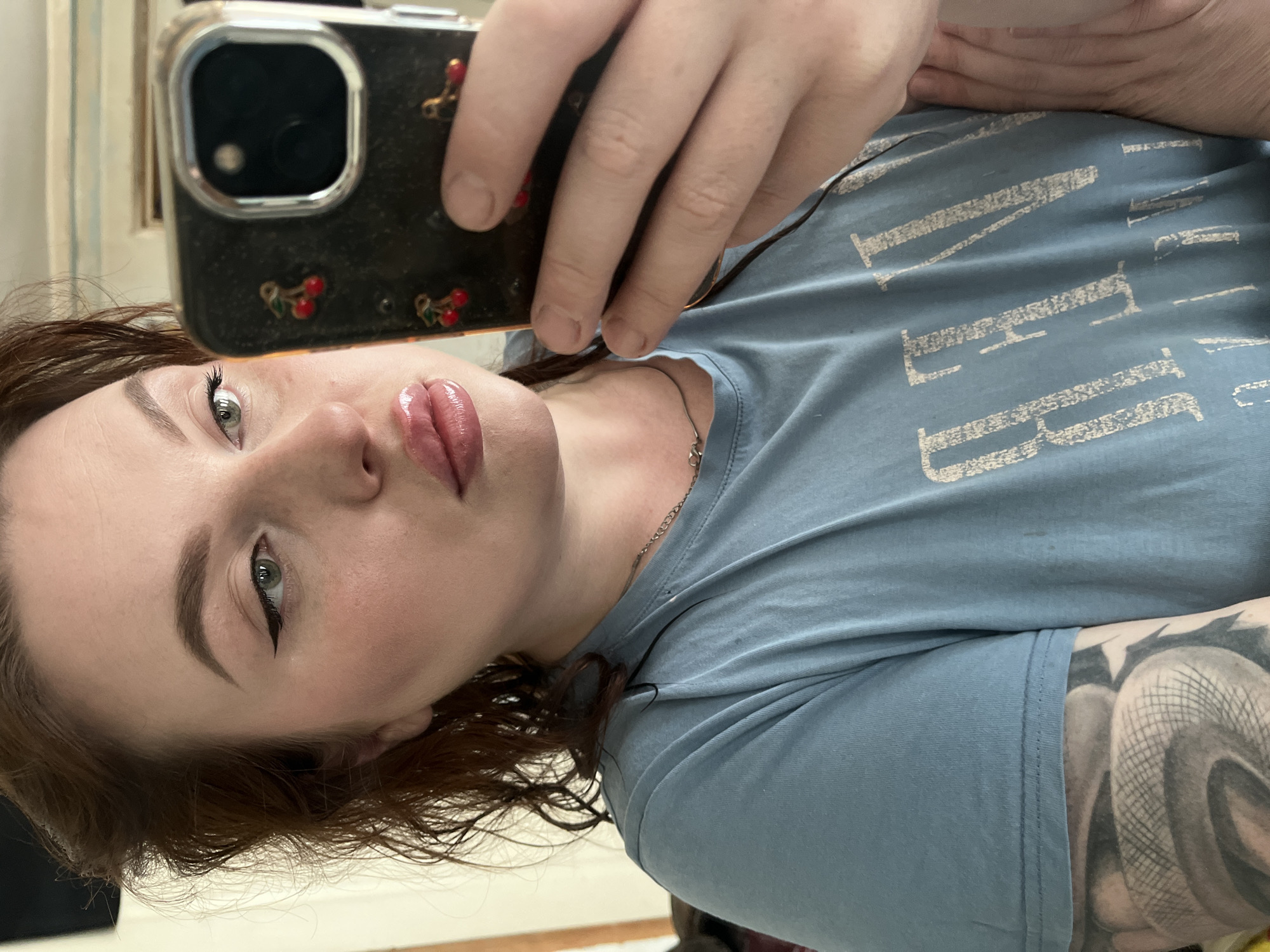 A fair-skinned woman with red hair is taking a selfie. She is wearing a light blue t-shirt with white writing. Her left arm is visible with a large tattoo. She has eyeliner and glossy lips. The phone she is using has a clear case with cherries on it. The location is not discernible from the image.