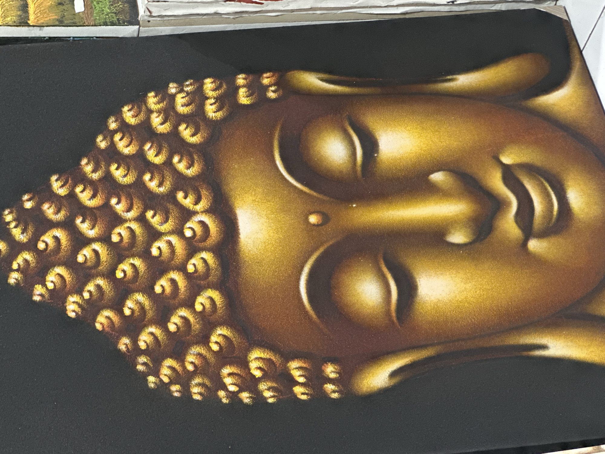 A golden Buddha head is depicted on a black background. The face is serene with closed eyes, a calm expression, and a smooth, textured golden finish. The head is adorned with stylized curls. The overall impression is that of a painting or a print displayed in Ubud District, Indonesia.