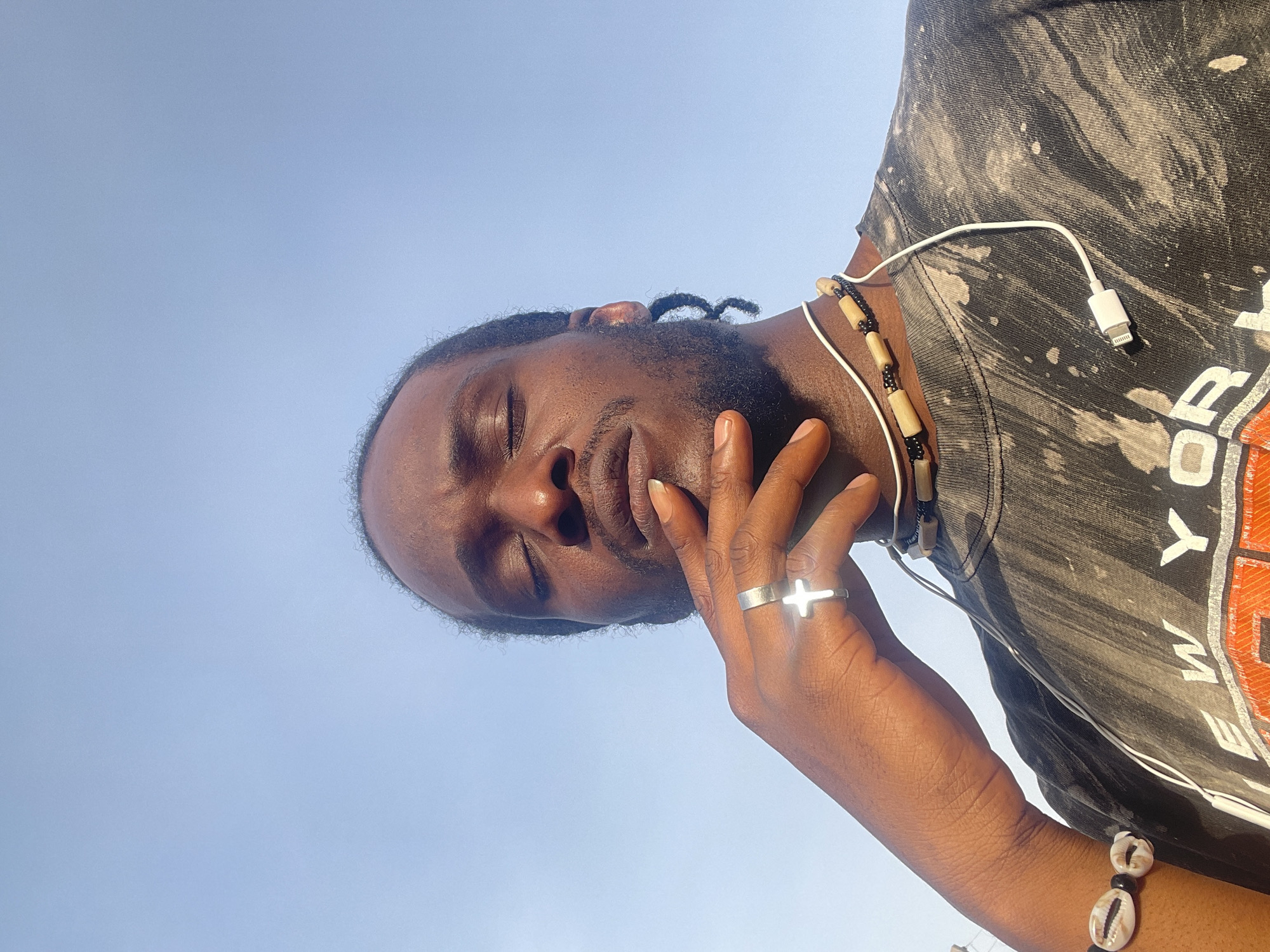 A low-angle, close-up shot shows a Black man with his eyes closed. He has short dreadlocks and a beard. His left hand is raised to his face, with a silver ring featuring a cross on his finger. He wears a dark-colored T-shirt with bleached patterns. A beaded necklace and a white earphone cable are visible around his neck. He also wears a beaded bracelet with cowrie shells on his wrist. The background is a clear, light blue sky. There is no indication of the location being Jos, Nigeria, based on the visual evidence provided.