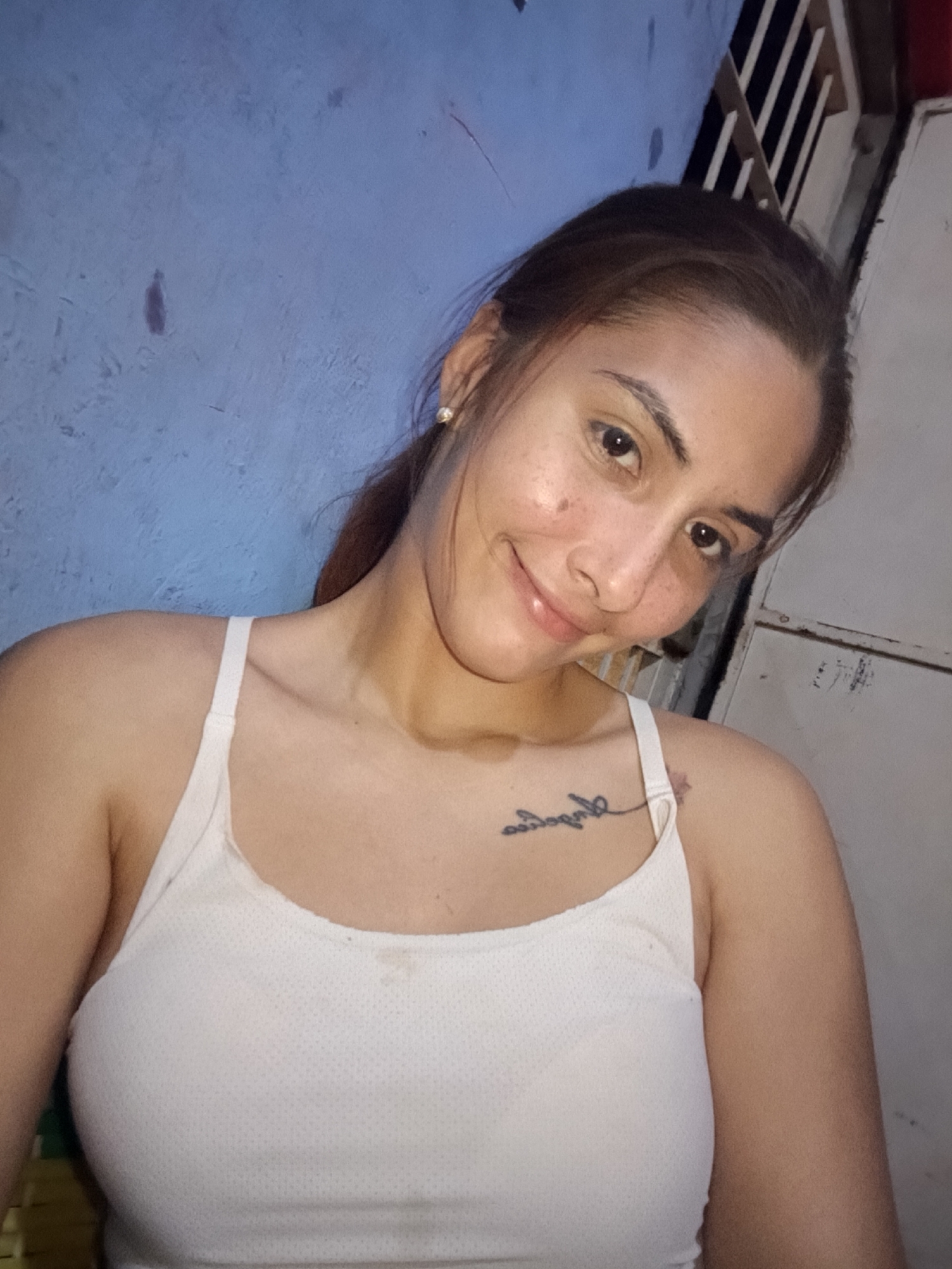 A woman with brown hair and light skin is centered in the image, wearing a white tank top and smiling slightly at the camera. She has a tattoo on her upper chest. The background includes a blue wall on the left and part of a building exterior with white or light-colored vertical slats on the right. There is no discernible context to associate this image with Juan Griego, Venezuela.
