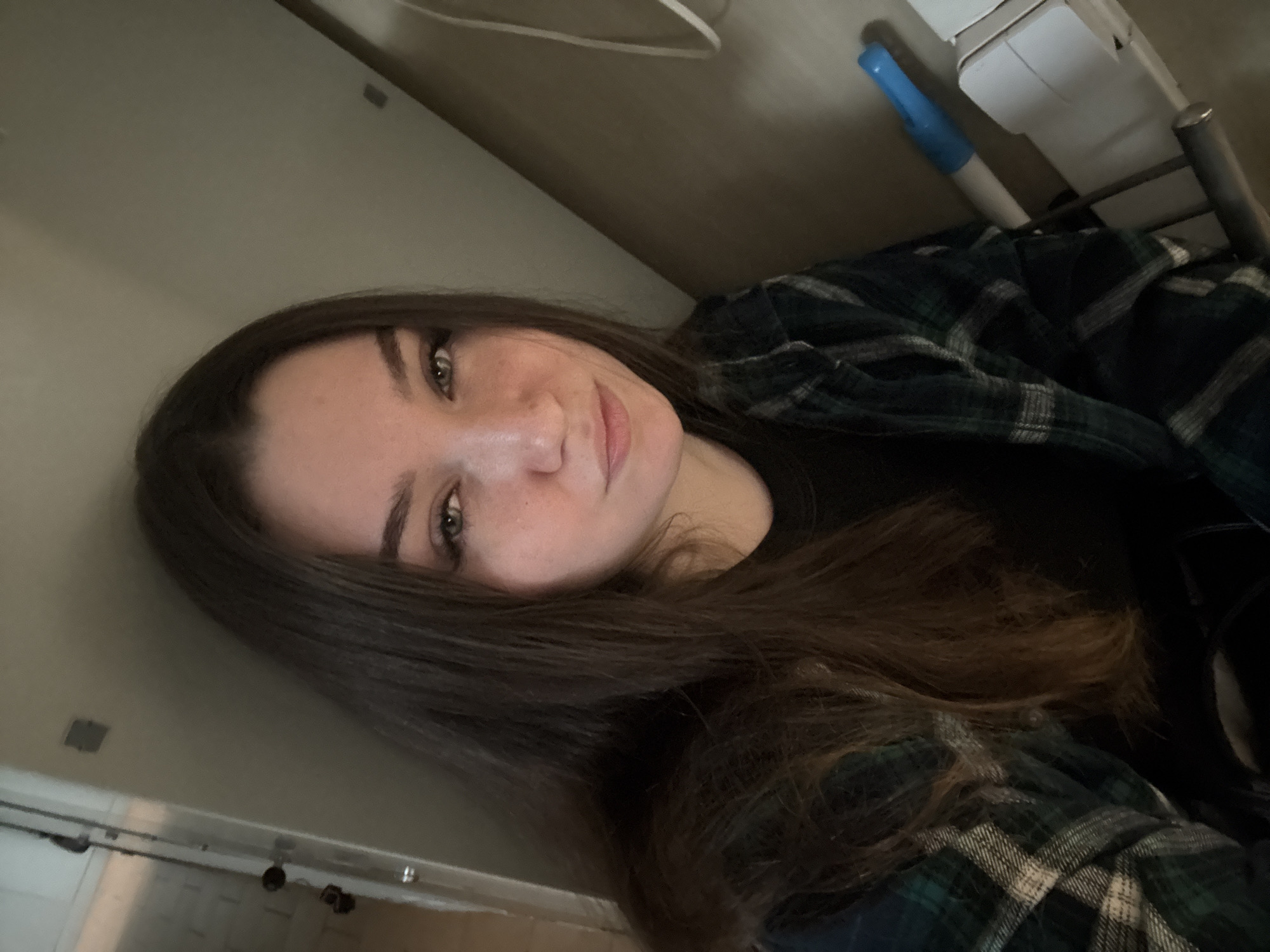 A young woman with long, dark brown hair and light green eyes is captured in a close-up, head-and-shoulders selfie. She is wearing a dark green and black plaid shirt over a black garment. Her head is tilted, and she is looking towards the camera with a slight smile. Light freckles are visible on her cheeks and nose. The background appears to be indoors, with a plain, light-colored wall behind her. To the upper right, a white shelf unit with a blue object, possibly a marker, and a metal rod is visible. The lighting suggests an indoor setting, possibly during the day. The overall mood is casual and relaxed. There is no visible text in the image. The location context of Сокол, Russia, is provided.
