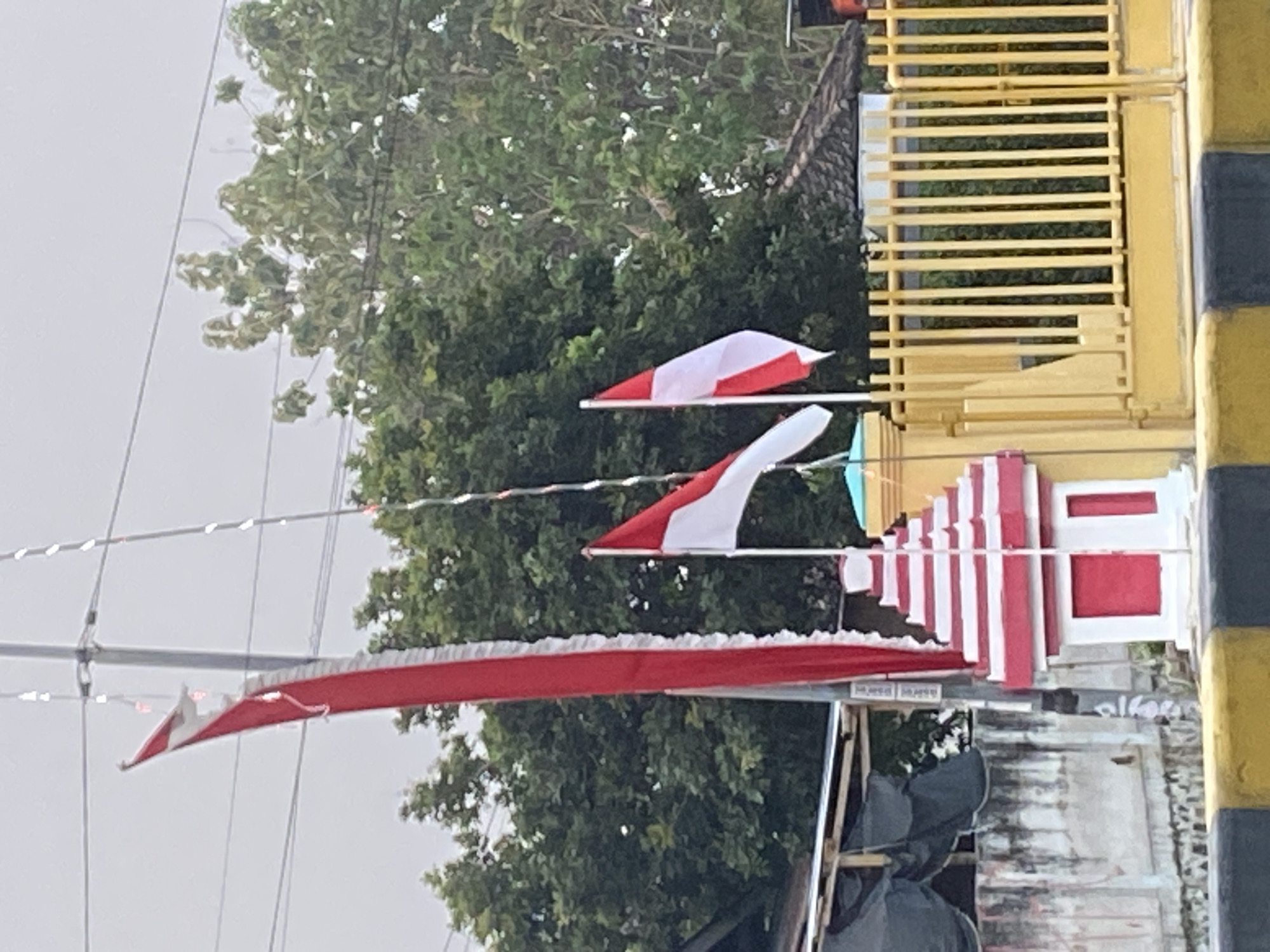 A street-level, medium-shot photograph captures a scene in Sugihwaras, Indonesia, featuring Indonesian flags, a decorative structure, and a yellow fence against a backdrop of trees and an overcast sky. Two Indonesian flags, red and white, are prominent. The flag on the right is fully visible, attached to a pole and waving. The flag on the left is a long, narrow red banner with a white frilled edge, partially obscured and hanging vertically. Next to the flags, to the left, is a small, multi-tiered, red and white decorative structure resembling a miniature pagoda or shrine. To the right of this structure is a bright yellow metal fence with vertical bars. In the background, dense green trees form a natural backdrop, with hints of buildings and utility poles visible. The foreground shows a road divider with black and yellow stripes. The overall impression is of a festive or commemorative display on a cloudy day.
