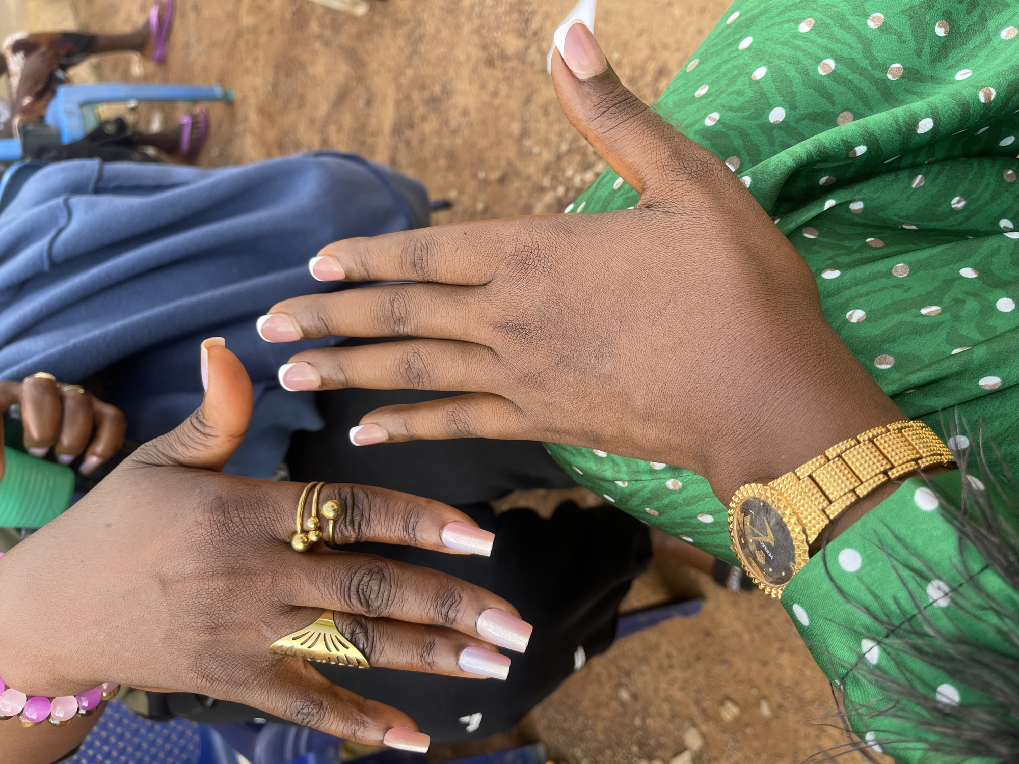 The image shows a close-up view of two hands with manicured nails and jewelry. The hand in the foreground is wearing a gold-toned watch with a black face and a textured band. It is also adorned with a green and white polka-dot dress. The second hand, positioned above and to the left, is adorned with two gold rings and a pink beaded bracelet. This hand has square-shaped nails painted in a pearlescent pink hue. The background is blurred, but appears to be an outdoor setting, possibly in Mangu Town, Nigeria, with hints of people and chairs. The overall lighting suggests it is daytime.
