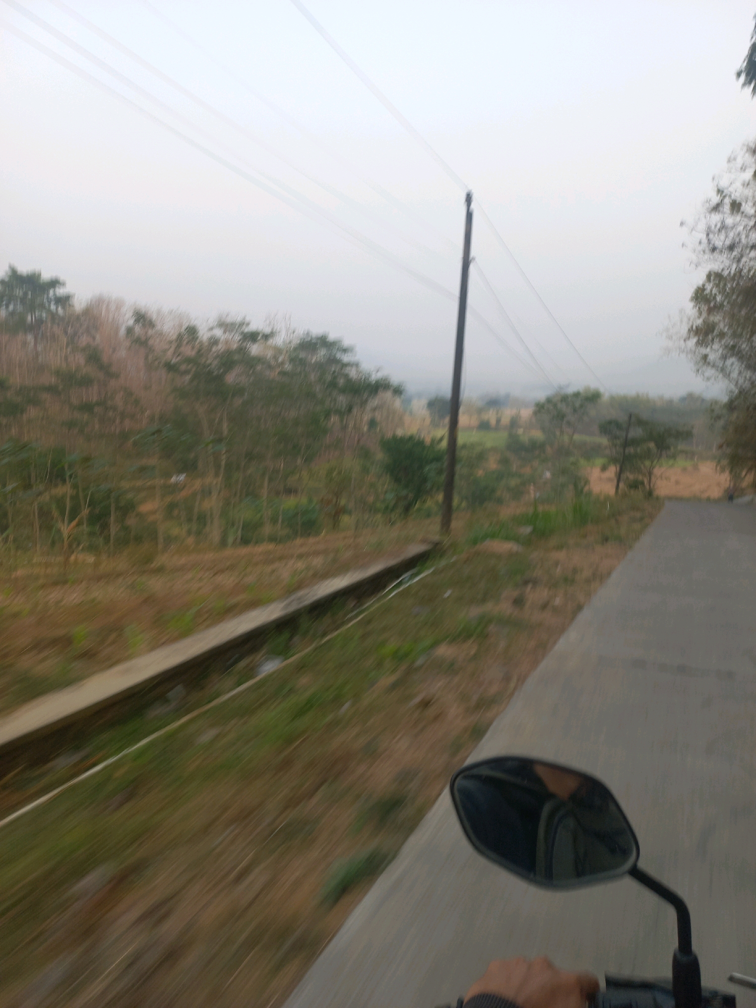 The image depicts a blurred view from a moving vehicle, likely a motorcycle, on a paved road. The scene is set in a rural or semi-rural area of Sukoharjo, Indonesia. On the left side of the road, there is a concrete drainage channel bordered by dry grass and some vegetation. Beyond the channel, a hillside covered with trees and brush is visible, appearing somewhat dry and sparse. In the distance, rolling hills under a hazy sky can be seen. A utility pole with power lines stretches across the frame. The reflection in the motorcycle's side mirror shows a person's hand, presumably the driver's, and a partial view of the driver's upper body. The motion blur suggests the photograph was taken while in transit.