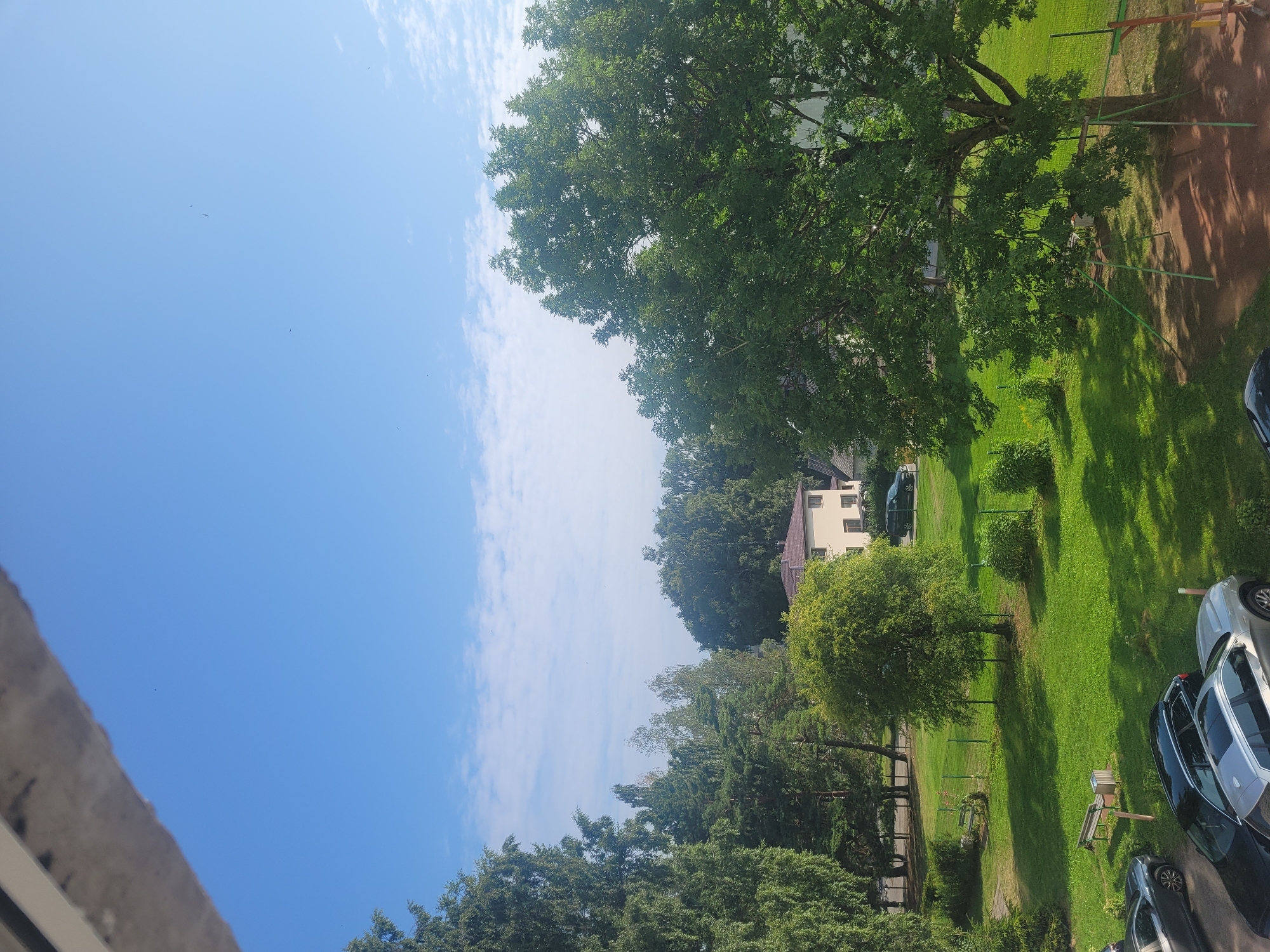 The image depicts an outdoor scene on a sunny day, likely in Jūrmala, Latvia, given the context. The foreground is dominated by the roof edge of a building, casting a shadow. Below this, a vibrant green lawn stretches across the middle ground, dotted with trees of varying sizes and types. In the background, a light-colored building with a brown roof is visible, surrounded by more trees. Several cars are parked on the lawn in the lower left portion of the image. A children's swing set is situated on the right side of the frame, partially in shadow. The sky above is a clear blue with scattered white clouds. The scene suggests a residential area with a park-like setting.