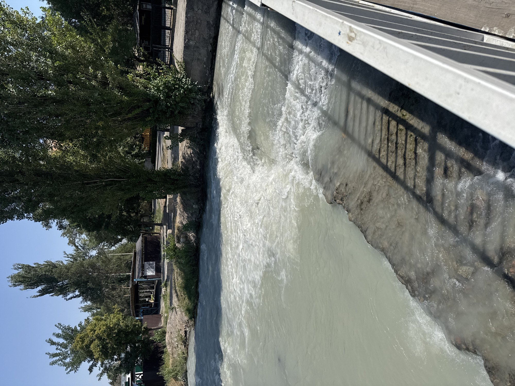 A natural, shallow river flows rapidly through an urban area under a clear blue sky. The water is a milky, light blue-grey color with visible white foam and eddies created by a small cascade. A concrete embankment lines the river, with some sections appearing eroded and covered in algae or sediment. On the left bank, a wooden fence and a small, dark-colored shelter with a corrugated roof are visible, surrounded by trees and greenery. On the right bank, a light-colored metal railing runs along the edge of a wooden walkway, casting a shadow of parallel bars onto the embankment and into the water. The background consists of more trees, some buildings, and a clear blue sky. The image appears to be taken from the walkway, looking downstream. This scene is consistent with the Alamedin River embankment in Bishkek, Kyrgyzstan, which is characterized by its natural flow through the city, urban infrastructure, and surrounding vegetation.