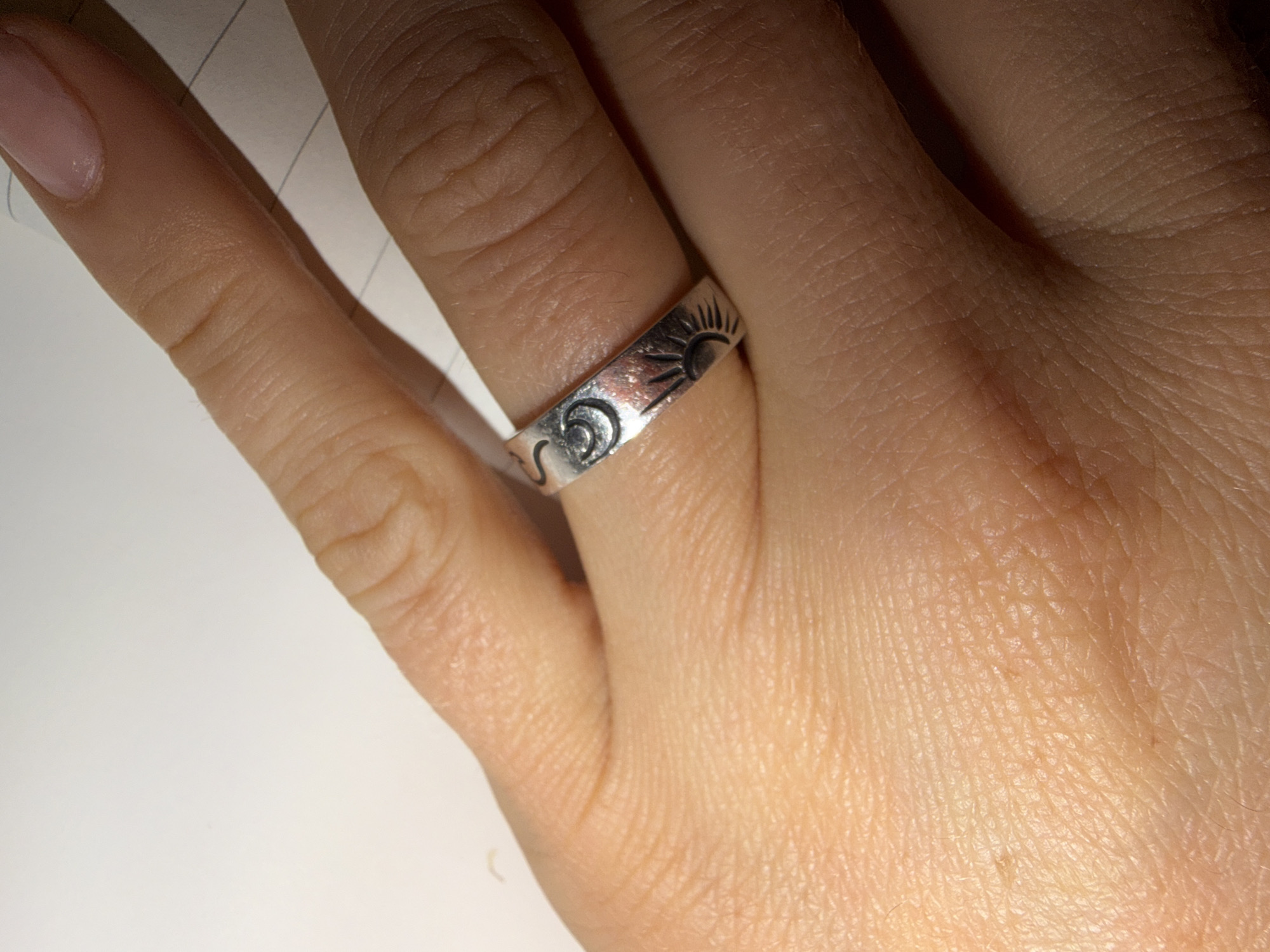 A close-up image shows a light-skinned hand wearing a silver ring with sun and moon designs. The ring is on the ring finger. The fingernail on the index finger is painted with a nude color. The background is plain and light. No location information is present.