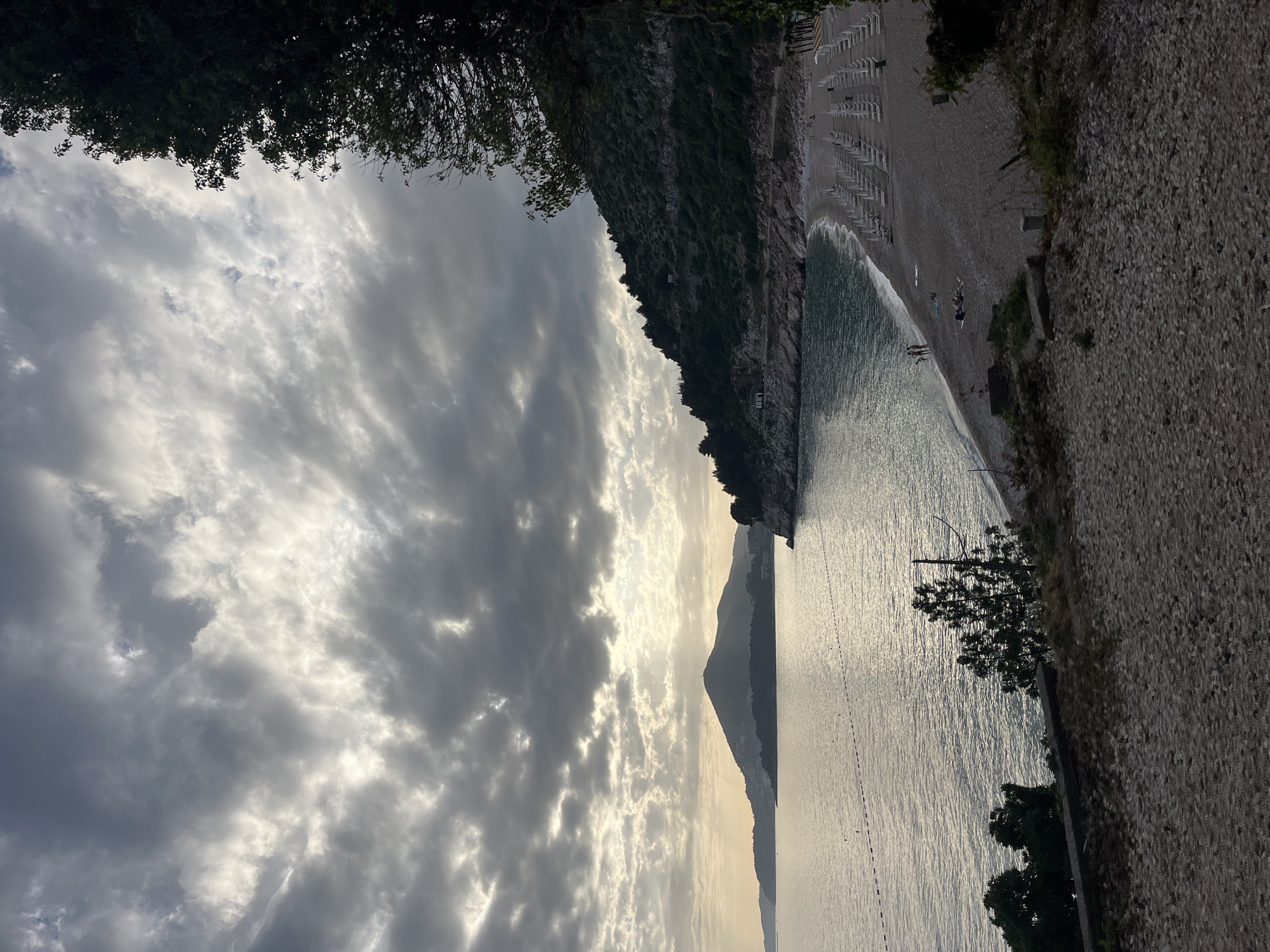 The image depicts a view of the Adriatic Sea meeting a beach in Bečići, Montenegro, under a sky filled with layered, gray, and white clouds. The sun appears to be behind the clouds, casting a diffused light on the water and sand. A steep, rocky cliff covered in vegetation rises on one side of the beach, and a stony wall is on the other. Beach chairs line the shore. A few figures are seen on the beach near the water's edge. A mountain can be seen in the distance across the water. Trees are in the foreground both at the top and bottom of the image.