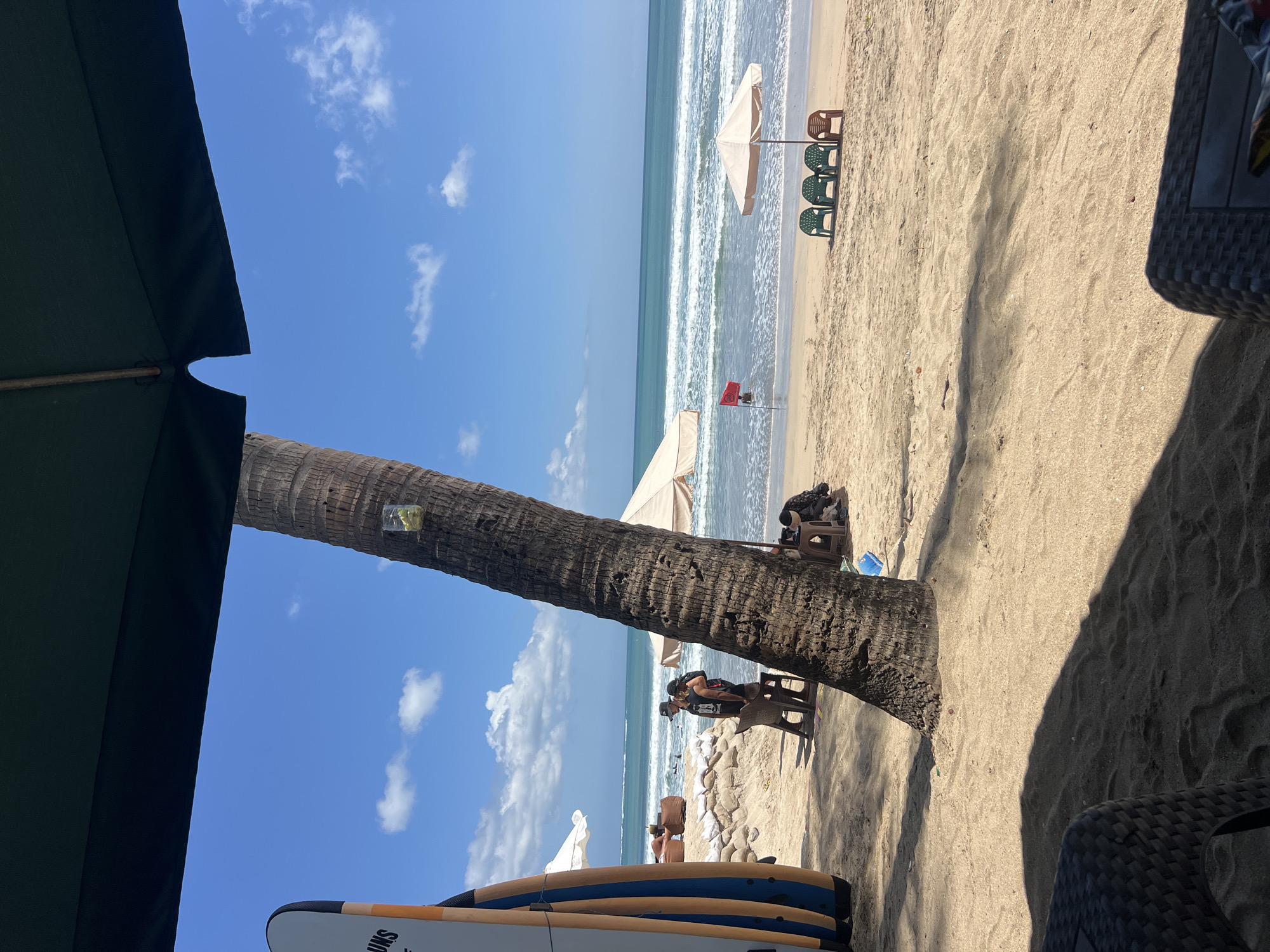 The image depicts a sunny beach scene in Kuta, Indonesia, with the ocean in the background and a prominent palm tree in the foreground. The beach is sandy, with some scattered debris. Several surfboards are leaning against a wall on the left side of the frame. In the middle ground, two people are standing near a table and chairs, and a red flag is visible in the water, indicating a potential surf break or warning. Further back, beach umbrellas and chairs are set up. The sky is blue with scattered white clouds. A dark-colored canopy extends over the top portion of the image, casting a shadow on the sand in the foreground. The overall impression is a relaxed beach environment with signs of recreational activity.