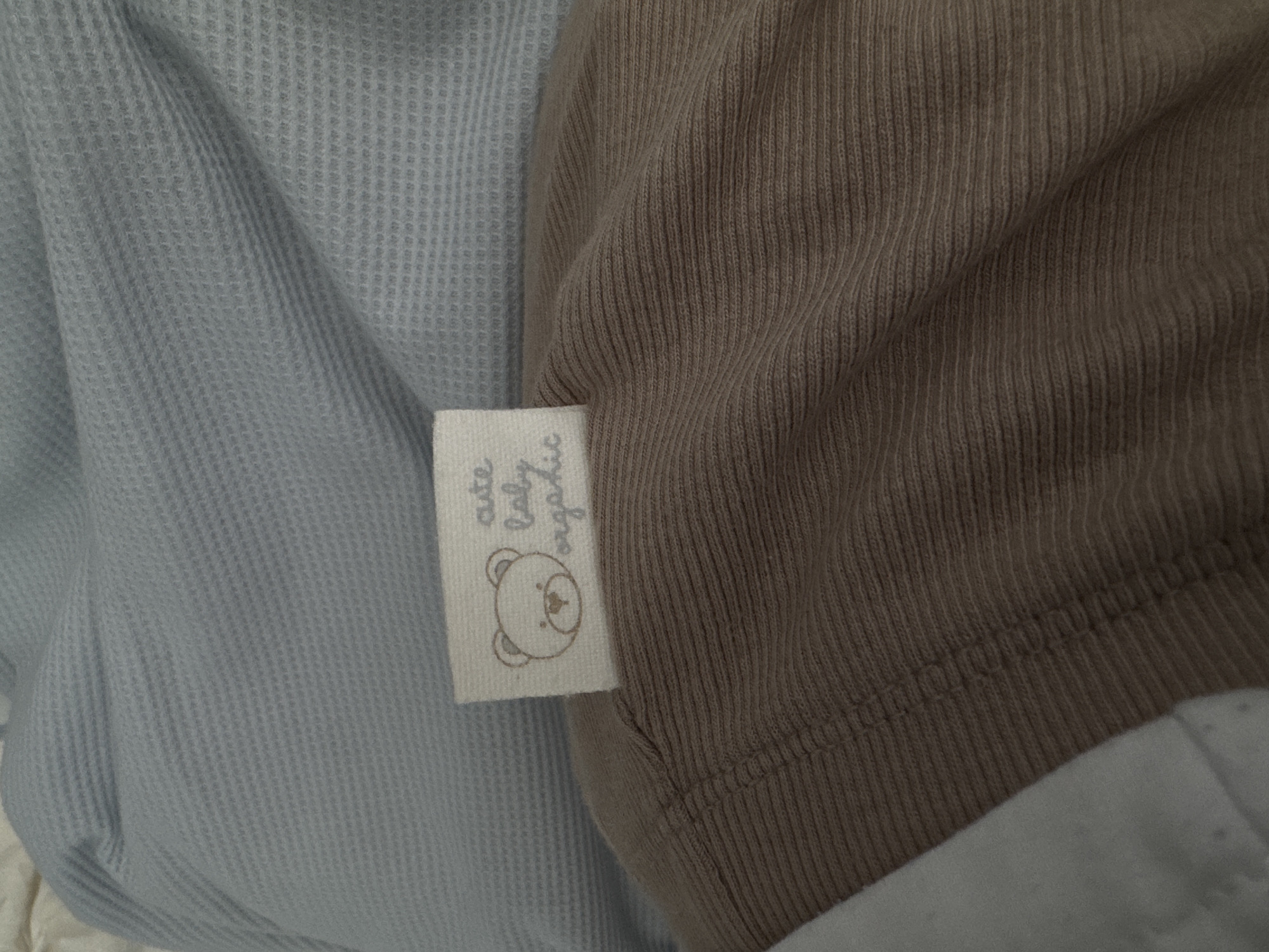 This is a close-up, eye-level photograph of a label attached to a piece of light brown, ribbed fabric. The label is a light beige rectangle with a simple line drawing of a teddy bear's face on the left side. To the right of the bear, text in a handwritten font reads "cute baby organic." The fabric appears to be a soft material, possibly cotton, and is draped over a light blue, waffle-knit fabric. The background is out of focus, showing the textures of the two fabrics. There is no indication of a specific location in Siegburg, Germany.