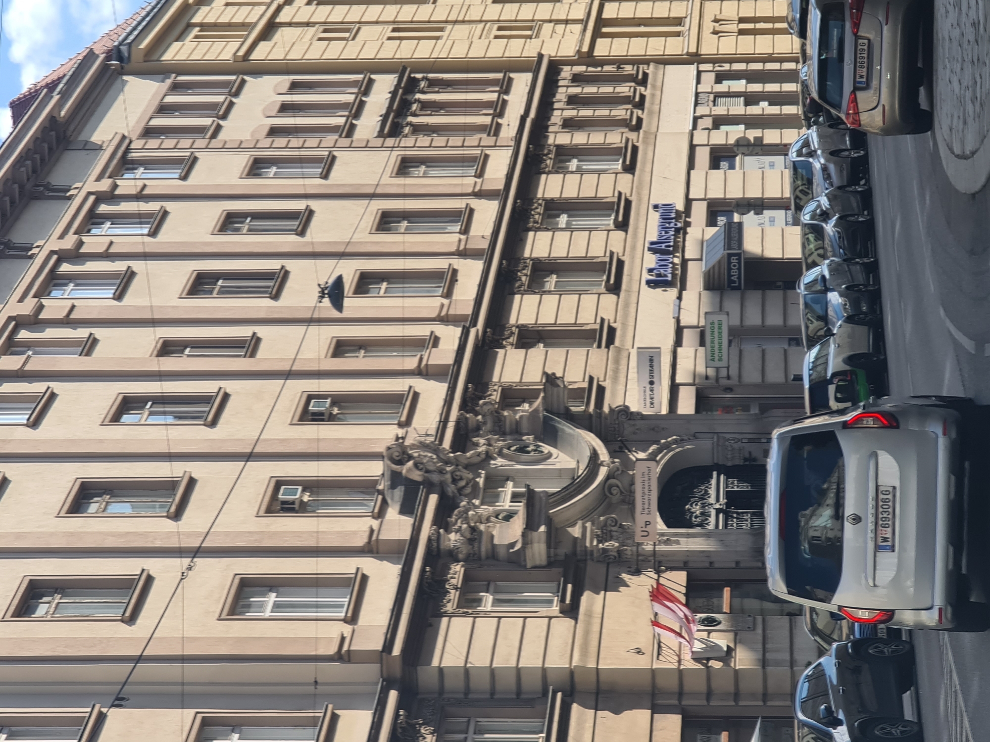 The image captures a street scene in Vienna, Austria, with a focus on a building facade and traffic. The building, characteristic of Viennese architecture, is a multi-story structure with a light beige facade, featuring numerous windows with white frames and ornate architectural details around the entrance. The entrance itself is grand, with decorative stonework and a curved overhang. Above the entrance, signs indicate businesses such as "Tierarztpraxis im Schwarzspanierhof" (Veterinary Practice in Schwarzspanierhof), "Labor Alsergrund," and "Änderungs-Schneiderei" (Alteration Tailor). A street lamp hangs from wires strung across the building.
In the foreground, several cars are visible on the street. The most prominent is a silver minivan with license plate "W W69306 G". Other cars, including a dark SUV, a gray sedan, and a gold hatchback with license plate "W W86919 G", are also present, indicating typical urban traffic. The cars are parked or moving along a paved street. The lighting suggests it is daytime, with shadows cast by the building and cars, indicating sunlight. The sky is partially visible at the top of the image and appears blue with some clouds. Red and white striped banners hang near the entrance of the building on the left side of the image.