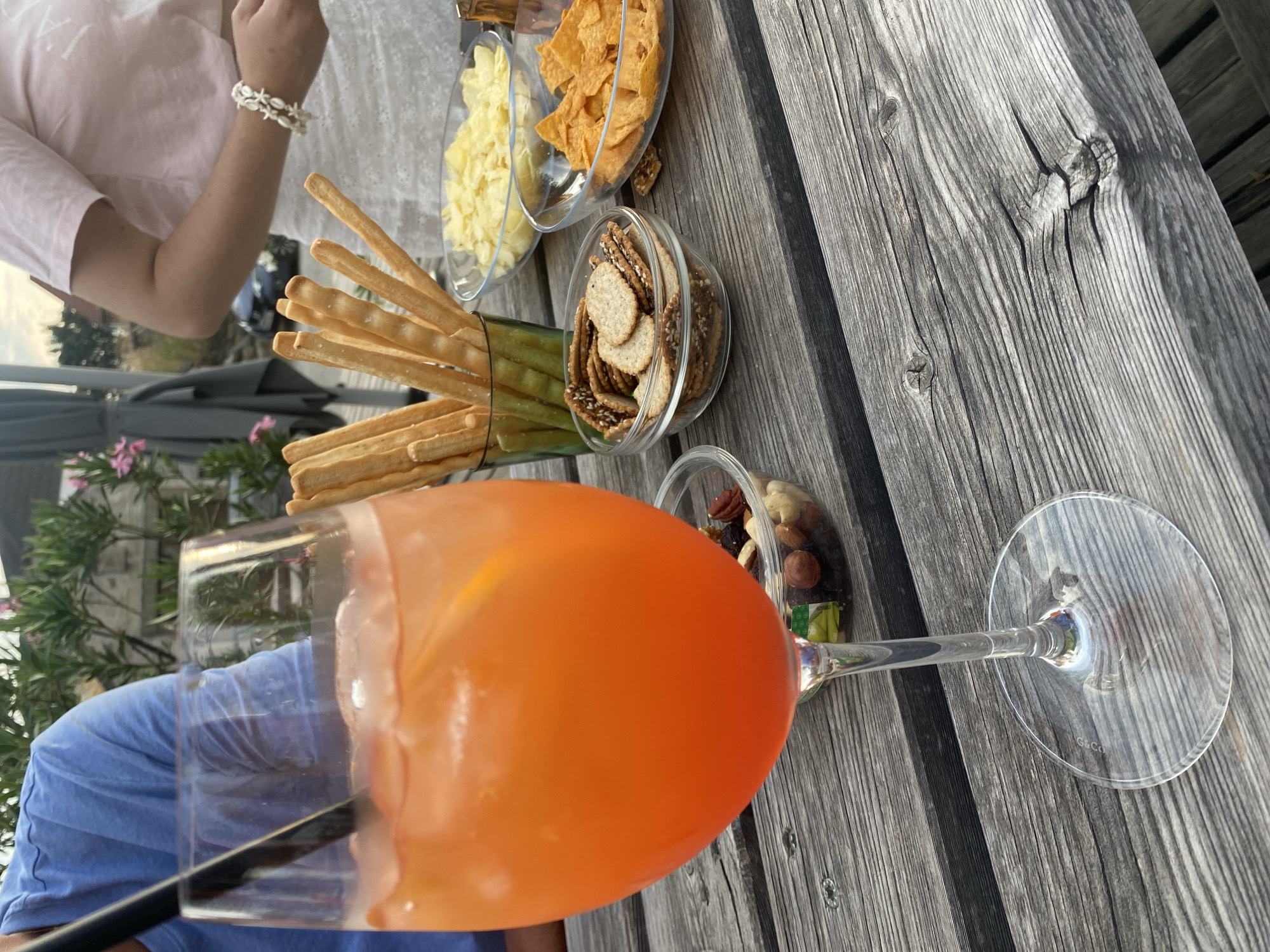 A close-up shot showcases a table laden with snacks and a vibrant orange cocktail. The cocktail, served in a large wine glass with a black straw and ice, is the focal point. Surrounding the drink are several bowls of snacks: breadsticks in a green glass, small crackers with sesame seeds, potato chips, and tortilla chips. Another small container holds a mix of nuts and dried fruit. In the background, two individuals are partially visible, one wearing a blue shirt and the other a pink top. The setting appears to be an outdoor seating area, likely a terrace or garden, with foliage and a car visible in the distance. The table itself is made of weathered wooden planks. The scene is located in Purbach, Austria.