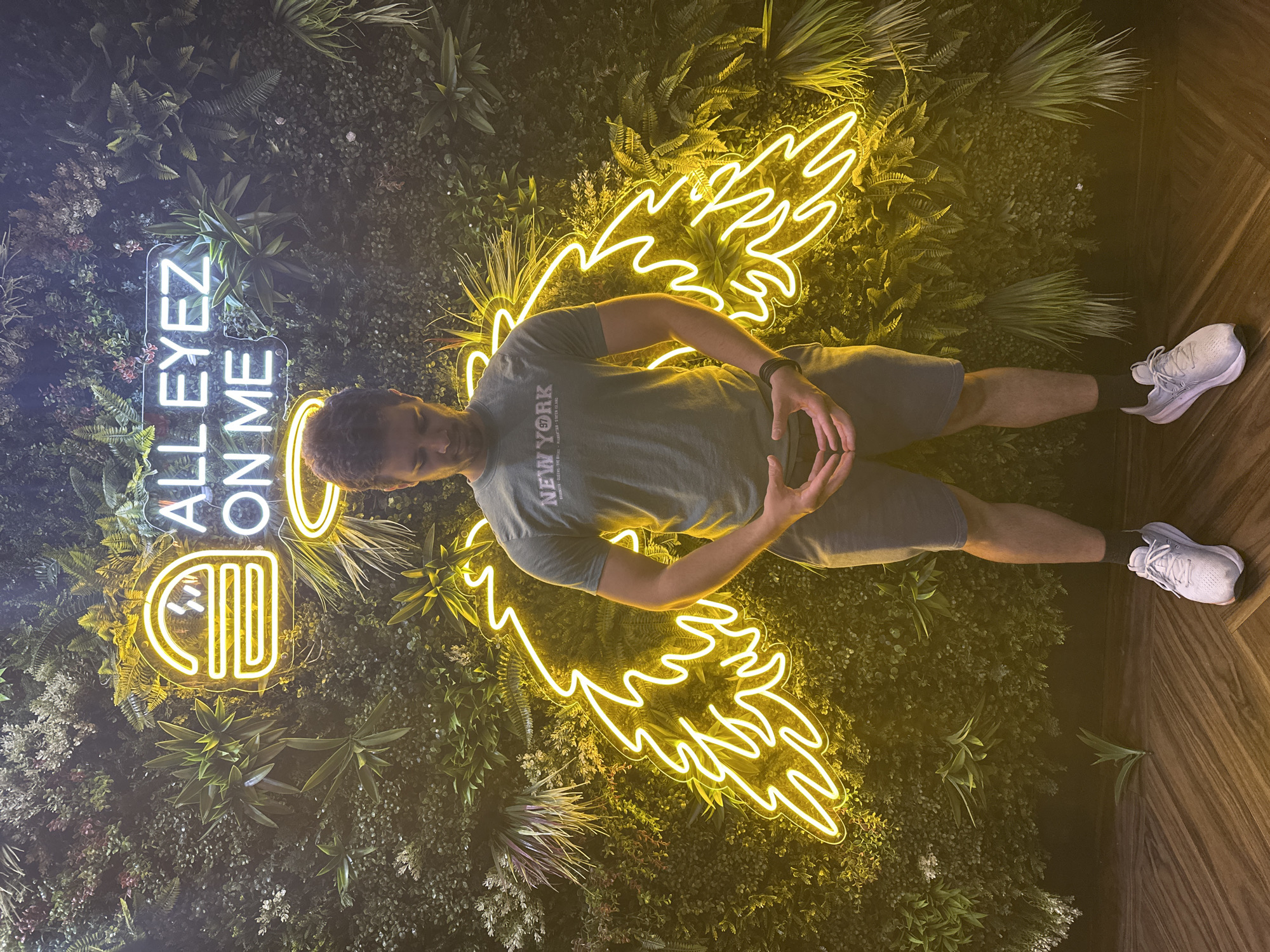 A young adult male stands in front of a vertical green wall adorned with various plants. Behind him, a neon yellow sign displays a burger icon and the text "ALL EYEZ ON ME." The neon light also forms a pair of angel wings behind the man. The man is wearing a grey t-shirt with "NEW YORK" printed on it, grey shorts, white sneakers, and black socks. He is looking down with his hands clasped in front of him. The floor is a dark wood with a plank pattern. This scene is captured in Leeds, United Kingdom.