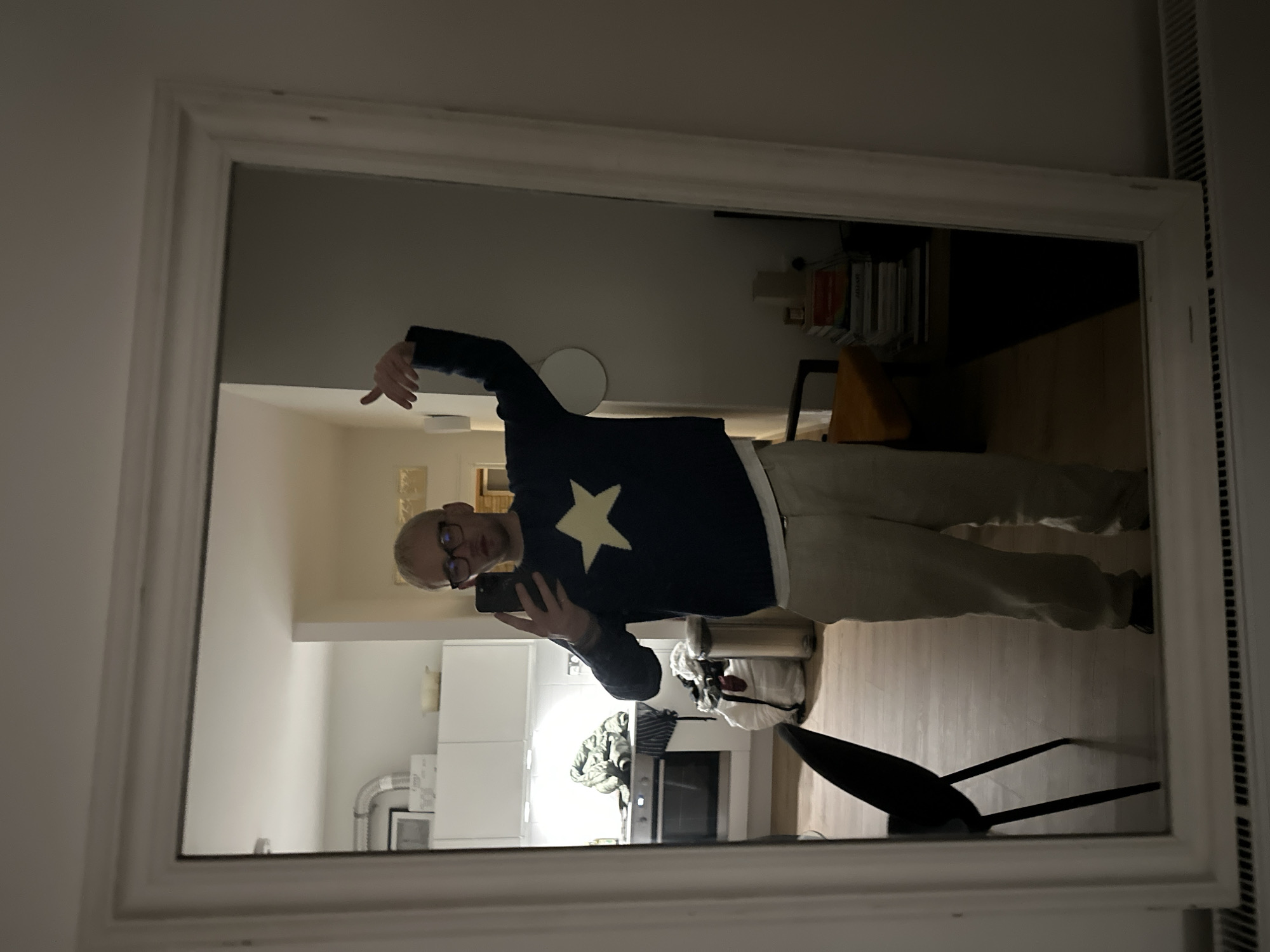 A person with short, light hair and glasses takes a selfie in a rectangular, white-framed mirror. The person wears a navy blue sweater with a large, yellow star and light-colored pants. The background reflected in the mirror includes a kitchen with white cabinets and stainless steel appliances. Behind the person is a room with books and a chair. A radiator is visible on the right. The scene appears to be indoors, likely in a flat or apartment, and cannot be identified specifically within the city of London, United Kingdom, from the given visual information.