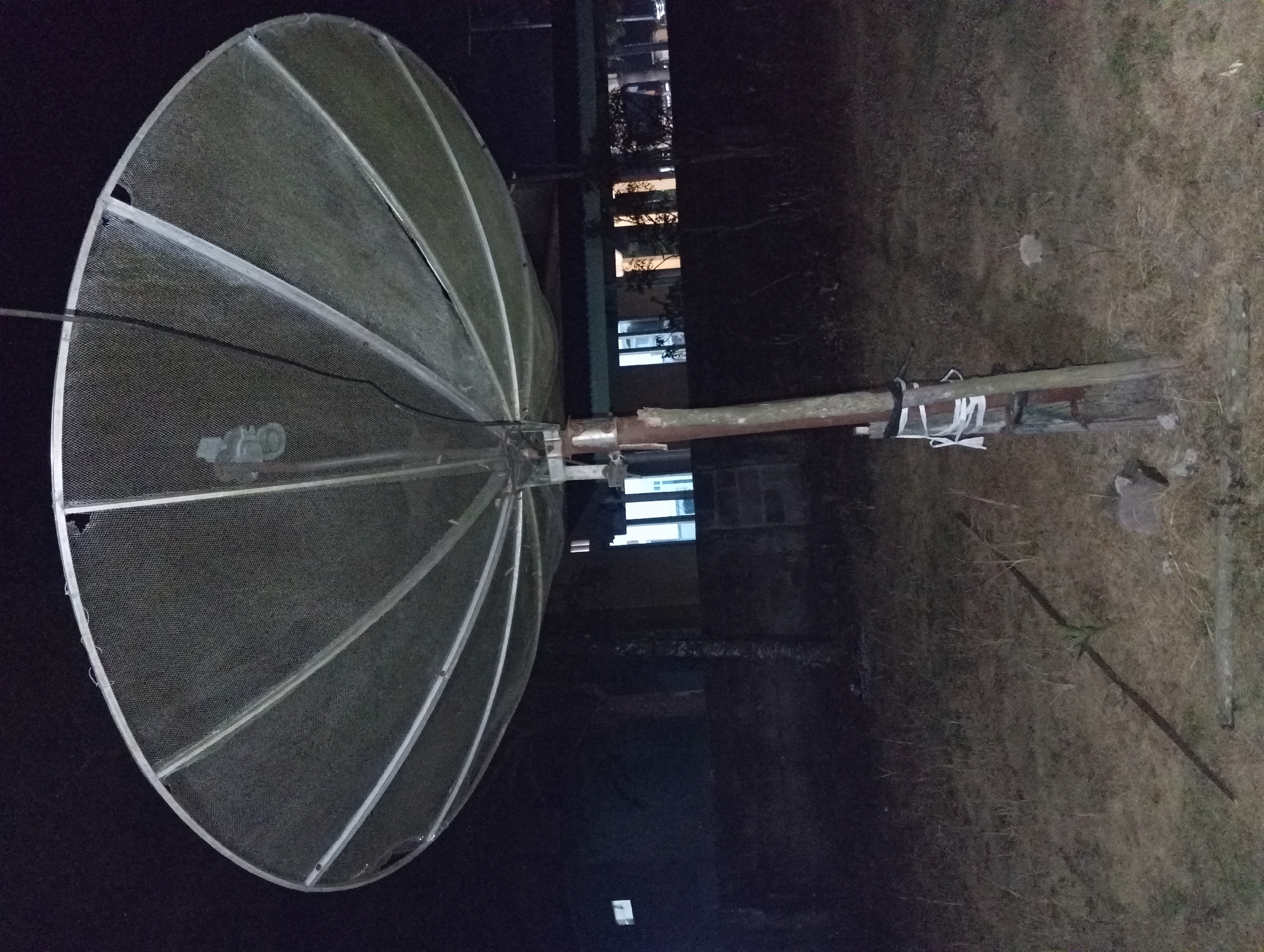 This image depicts a large, metal mesh satellite dish mounted on a weathered wooden pole at night. The dish is angled upwards, its surface supported by a radial metal framework. A black cable extends from the dish. The pole is secured with white and black strapping. In the background, a dimly lit building with illuminated windows is visible, suggesting a residential or commercial area. The ground in the foreground is covered with dry grass and scattered debris, including rocks and a piece of metal piping. The overall scene is dark, with the primary light sources being the building's windows and potentially ambient light from the surroundings. The location is specified as Rejosari, Indonesia.