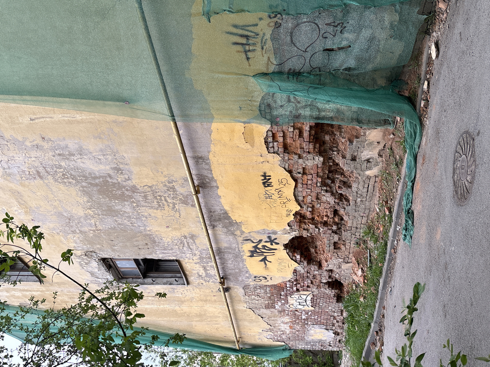 An exterior shot of a damaged building in Saint Petersburg, Russia. The building facade is a mix of aging stucco in beige and yellow tones, marred by peeling layers, graffiti, and significant structural damage exposing the red brick underneath. A green protective netting partially covers the building, seemingly to contain debris from the decaying structure. Two dark, recessed windows are visible on the facade. A metal pipe runs vertically along the wall. Adjacent to the building is an asphalt road with a concrete curb. A round manhole cover is visible on the road. Vegetation grows both in front of the building and alongside the road. The photo has been rotated 90 degrees, putting the road on the right side and the top of the building on the left.
