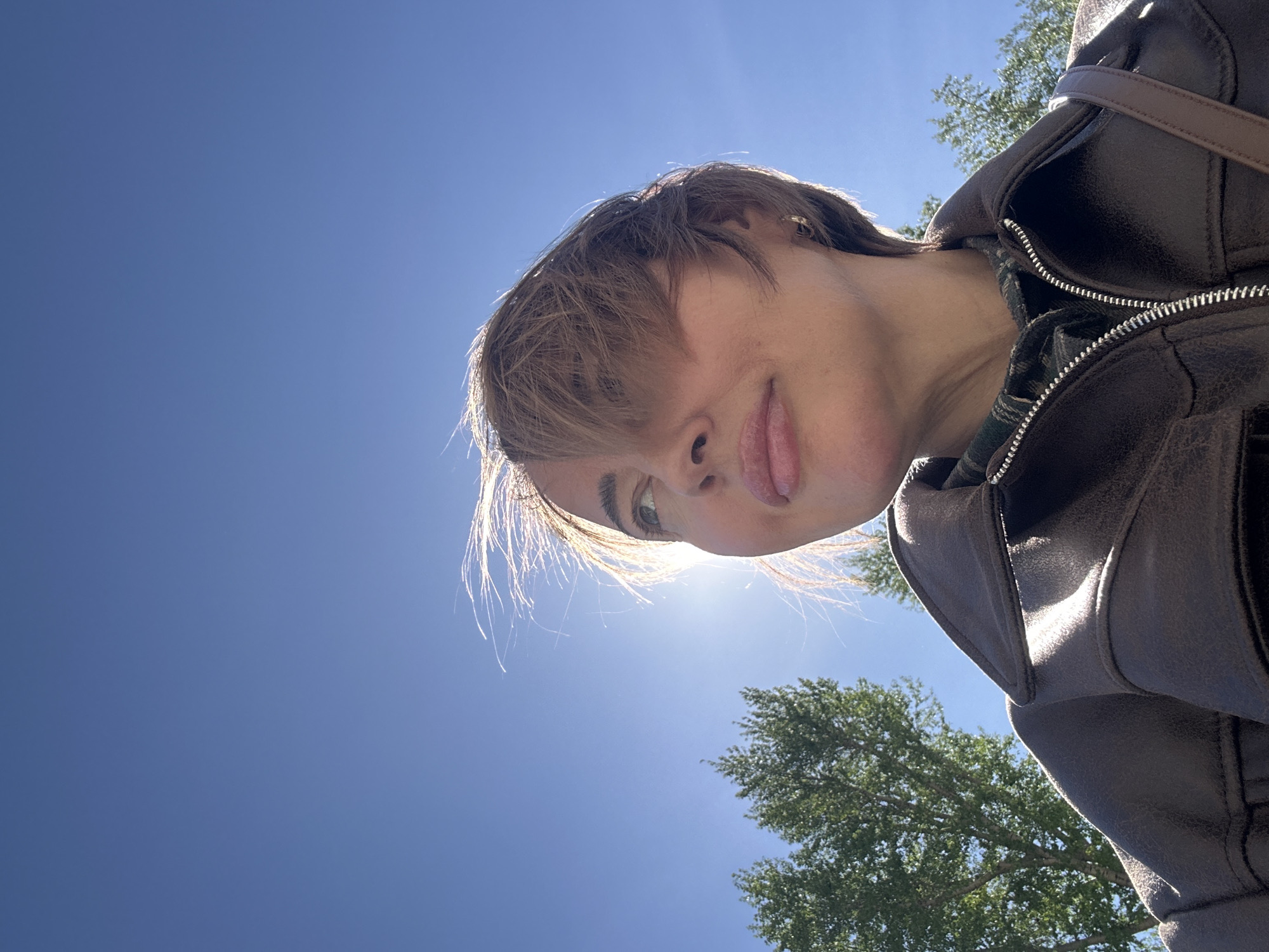 A low-angle photograph shows a person with short, light brown hair wearing a dark brown leather jacket. The person's face is visible from the nose upward, with their tongue slightly protruding. The background consists of a clear blue sky and the tops of green trees. No information is available to assess the location.
