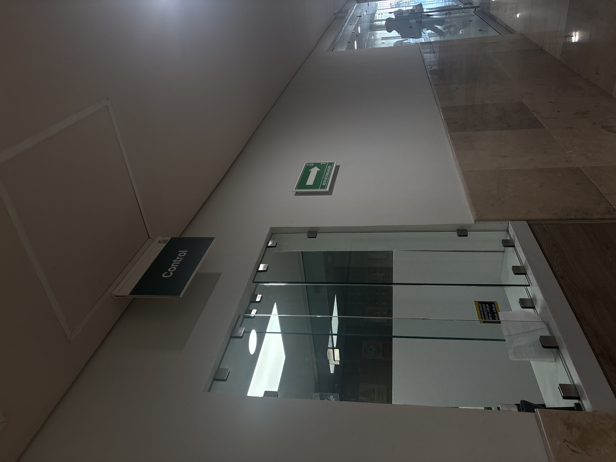 Here is a detailed description of the image:
**Setting:**
The image is taken in what appears to be a modern commercial building or clinic in Puebla, Mexico. The interior features marble flooring and minimalist white walls.
**Specific Features:**
* **Control Sign:** A sign labeled "Control" is mounted horizontally on the wall.
* **Glass Windows:** Below the "Control" sign are a series of glass windows, possibly indicating a reception area or service window.
* **Signage:** On the glass windows, there is a sign with the text "RARIO DE ATENCION TREGA DE ESTUDIOS 1:00 am-1:00 pm 4:00 pm-6:00 pm."
* **Evacuation Route Sign:** A green square sign with a white arrow and the words "RUTA DE EVACUACIÓN" (Evacuation Route) is visible on the wall, pointing in a specific direction.
**People and Activity:**
* The image captures a person passing by the glass windows, reflected in the glass.
**Environment:**
* The area seems well-lit. The minimalist design gives a clean and professional feel to the area.