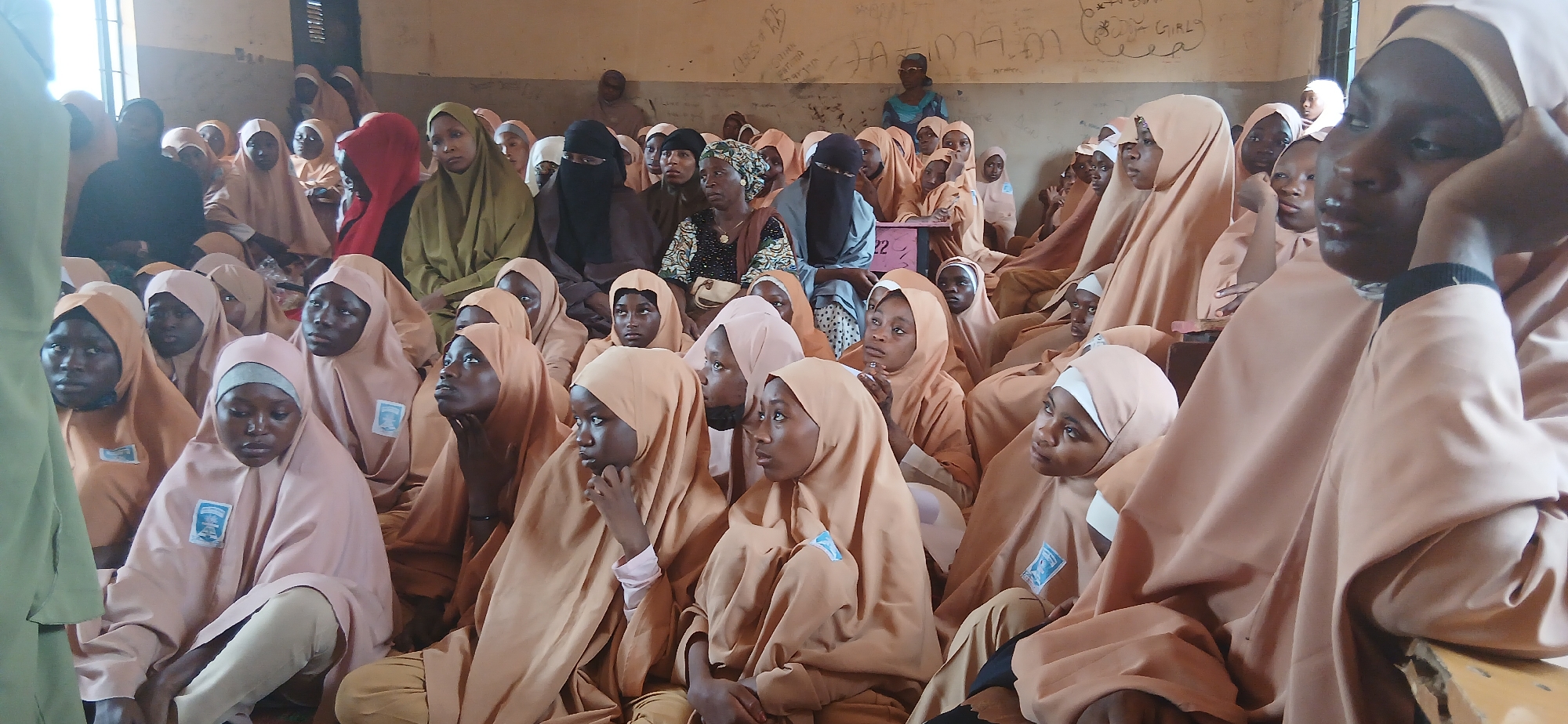 In an interior scene in Hayin Banki, Nigeria, a group of young people, primarily girls, are seated on the floor and on a bench. They are mostly wearing light brown or peach-colored hijabs and uniforms, with some wearing dark headscarves and outfits. The environment appears to be a classroom or assembly hall, with a wall in the background that is covered in writing and graffiti. There are windows on the left side of the image providing natural light. The individuals are facing forward, suggesting they are listening to someone or observing something outside the frame. The atmosphere is one of attentiveness, with some individuals looking directly at the viewer.