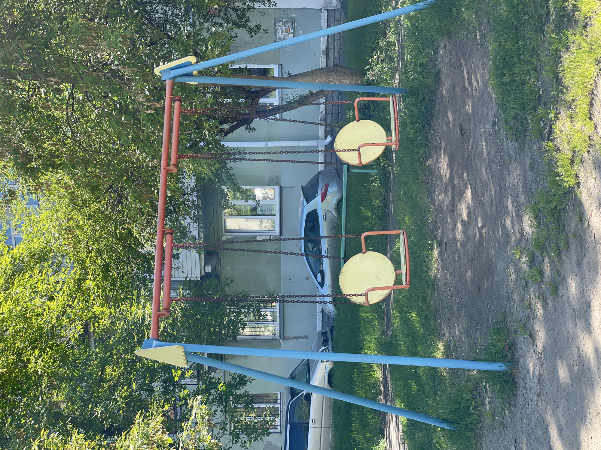 The image displays an empty children's playground in Zheleznogorsk, Russia, captured on a sunny day. In the foreground, a metal swing set dominates the scene, featuring a blue frame with orange-red support beams. Two yellow, circular swings hang from metal chains.
Behind the swing set, a light green or grey residential building stands, characterized by multiple windows and a white downspout. Two cars are parked near the building: a light-colored hatchback or sedan is clearly visible, and part of a darker vehicle can be seen further along. Large, leafy green trees provide a lush backdrop and frame the left side of the image, while grass and a well-trodden dirt path make up the ground around the playground.
Notable details include bright sunlight casting shadows on the dirt path. On the wall of the residential building, near the parked car, the text "K 5" is visible, along with a small, unreadable rectangular plaque above it. The scene is devoid of people, giving it a quiet, calm atmosphere under clear skies.