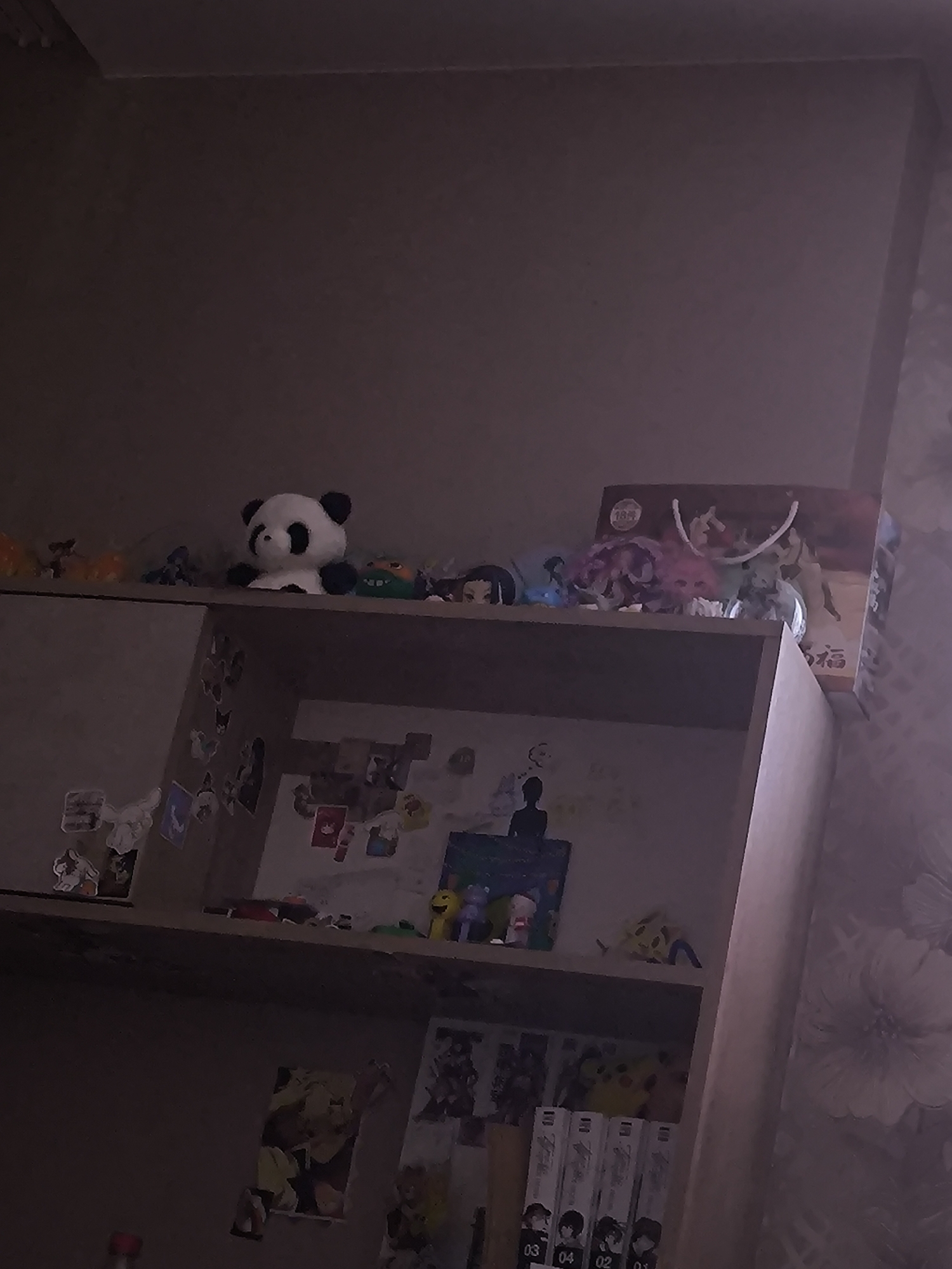 The image shows a dimly lit shelf filled with various collectible items. A stuffed panda bear sits prominently on the top shelf, accompanied by several anime figurines and what appears to be a gift bag with Japanese writing. The shelves below are also decorated with stickers and more figurines, including a silhouette of a person. The scene is set indoors, likely in a residential room. There are no people present in the image. The overall impression is of a personal display of cherished items. The image location is not discernible.