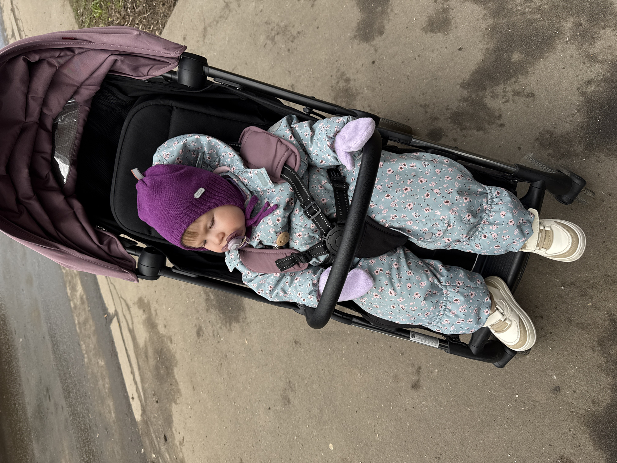A baby is asleep in a black stroller with a partially collapsed purple sunshade on a gray concrete surface, likely a sidewalk. The baby is wearing a purple knit hat, a light blue floral patterned snowsuit with purple mittens, and white sneakers. The stroller's safety straps are fastened across the baby's chest. The location may be Moscow, Russia.