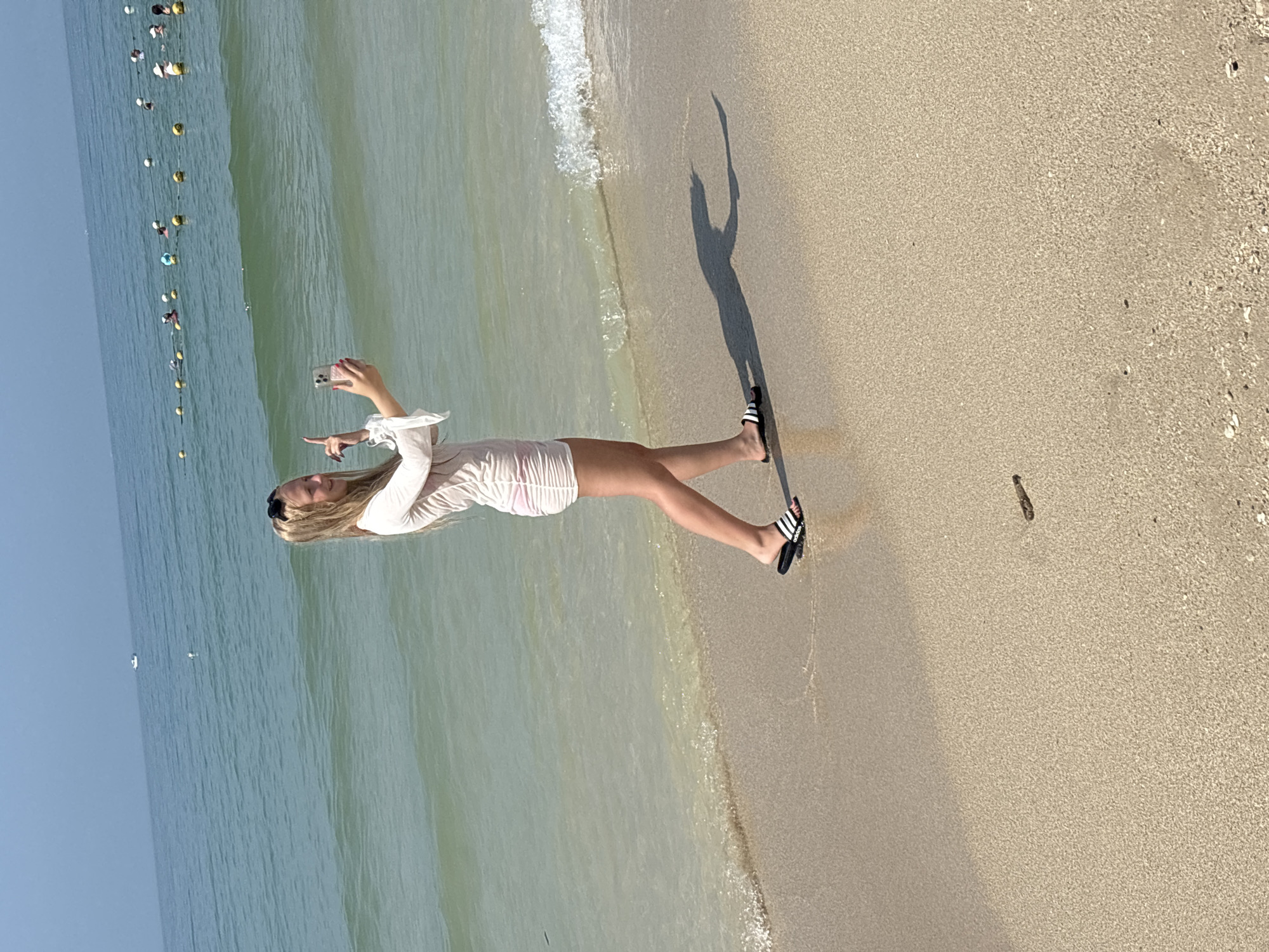 A young woman with blonde hair is standing on a sandy beach, facing the ocean and taking a selfie with her phone. She is wearing a light-colored, long-sleeved cover-up over a pink and white striped outfit, and black slides with white stripes. The ocean is calm with small waves breaking on the shore, and buoys with yellow and white markings are visible in the distance, suggesting a designated swimming area. Several people are visible in the water near the buoys. The sky is clear and blue. The overall scene depicts a leisurely day at the beach. The location is Jiyang District, China.