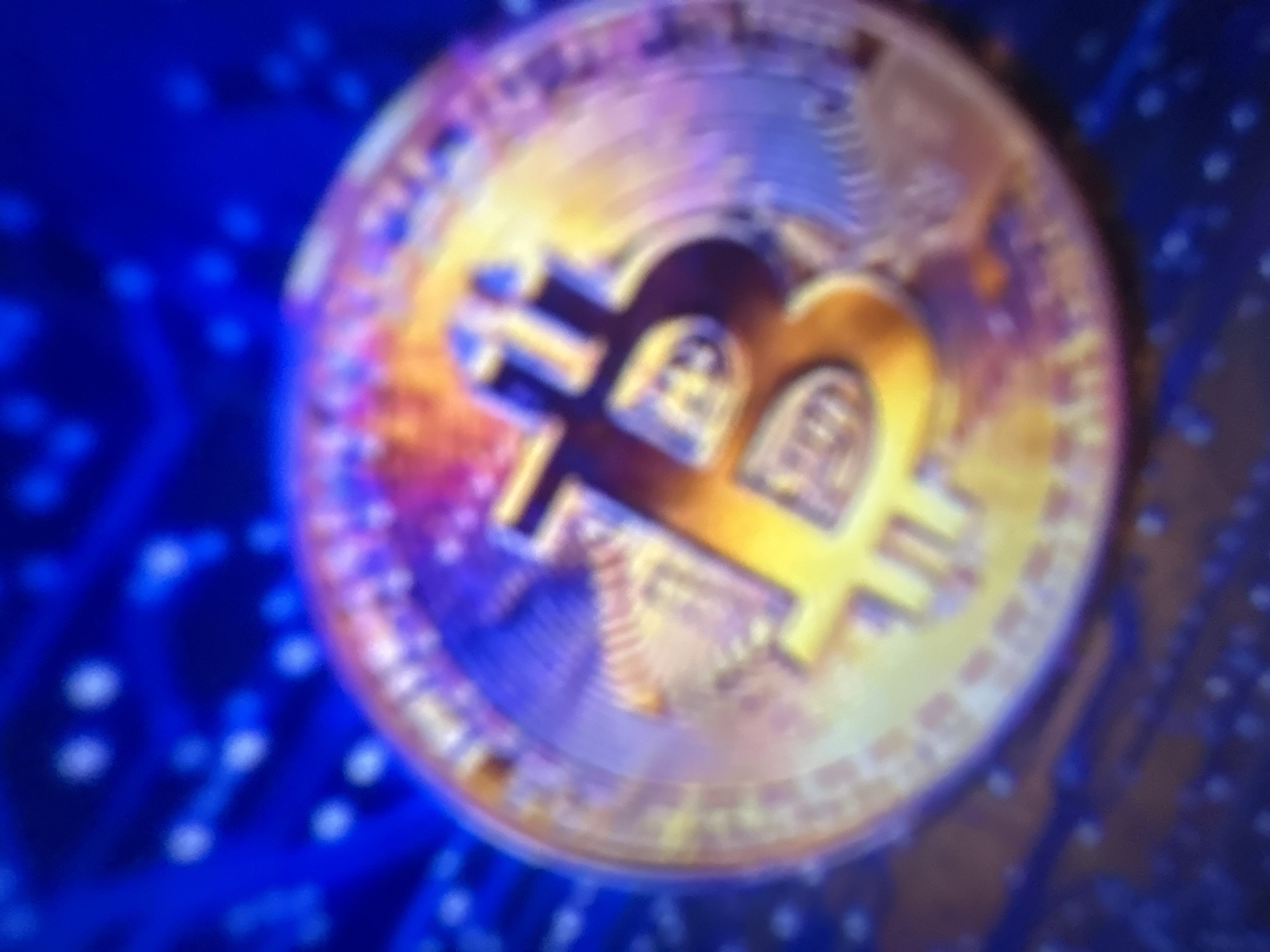 The image is a close-up, out-of-focus shot of a Bitcoin coin. The coin is gold-colored and features the Bitcoin symbol. It is placed against a blurred blue background with some glowing circular patterns, resembling circuitry or a digital landscape. The lighting is vibrant, with purple and yellow hues reflecting off the coin. The image appears to be taken indoors, with no visible elements of the city of Gelendzhik, Russia.
