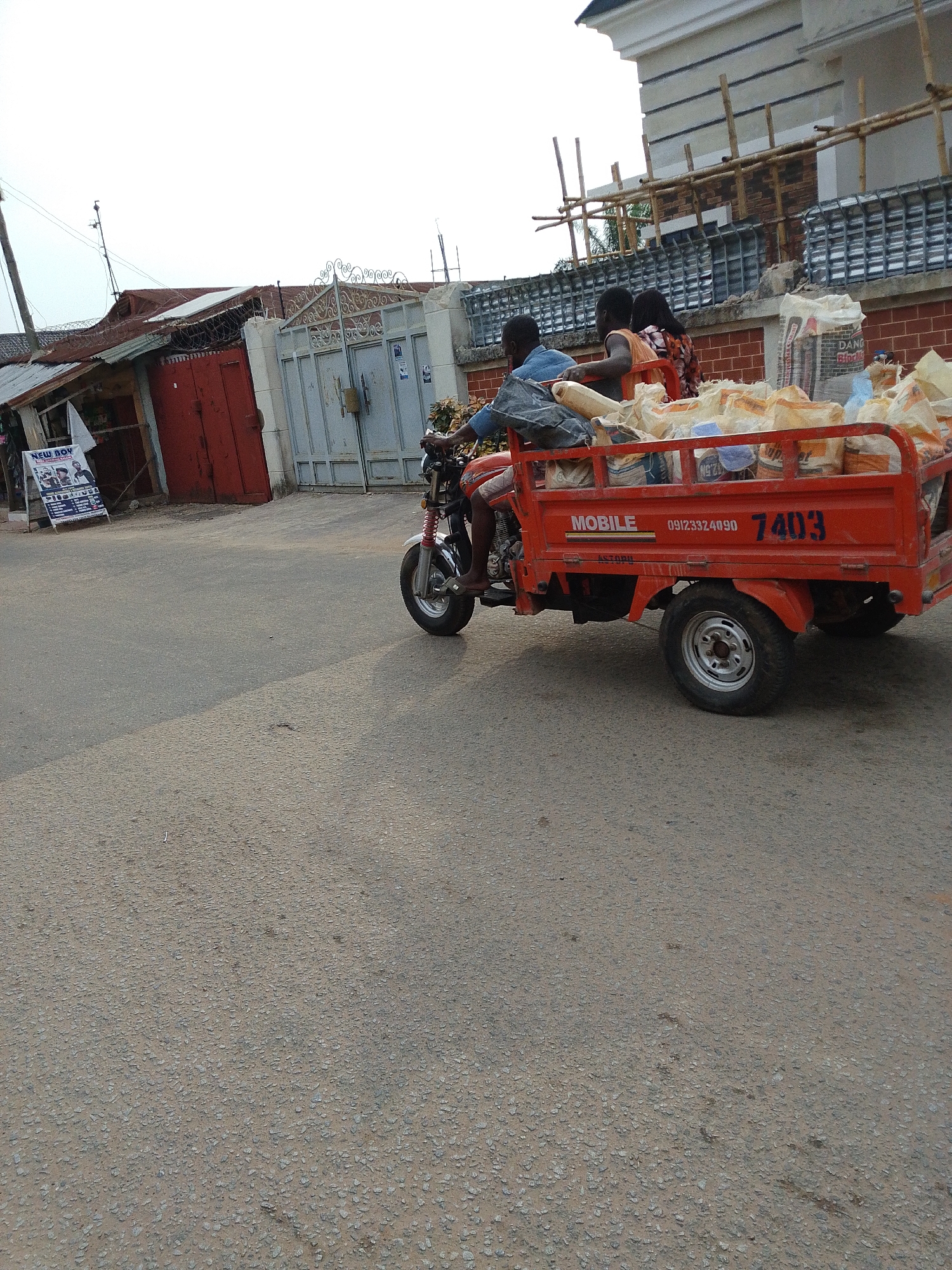 The image captures a motorized tricycle in motion on a paved road. The tricycle is painted red, with the word "MOBILE," a phone number (09123324090), and the number "7403" prominently displayed on its side. The tricycle is loaded with bags, possibly cement, and carries three individuals: a driver and two passengers. The driver is wearing a blue shirt, while the other passengers wear casual clothing.
In the background, buildings and structures are visible. To the left, there is a small, possibly commercial building with a red facade. Adjacent to this building is a sign advertising a barber shop called "NEW BOY." Further in the background, there is a gate and the facade of a larger building, possibly a residence, which is under construction as evidenced by the bamboo scaffolding erected around the building.
The image was captured during daylight hours, under an overcast sky, and the location appears to be Ohokobe Ndume, Nigeria. The scene suggests a typical urban environment in the region, with commercial activity and ongoing construction present.
