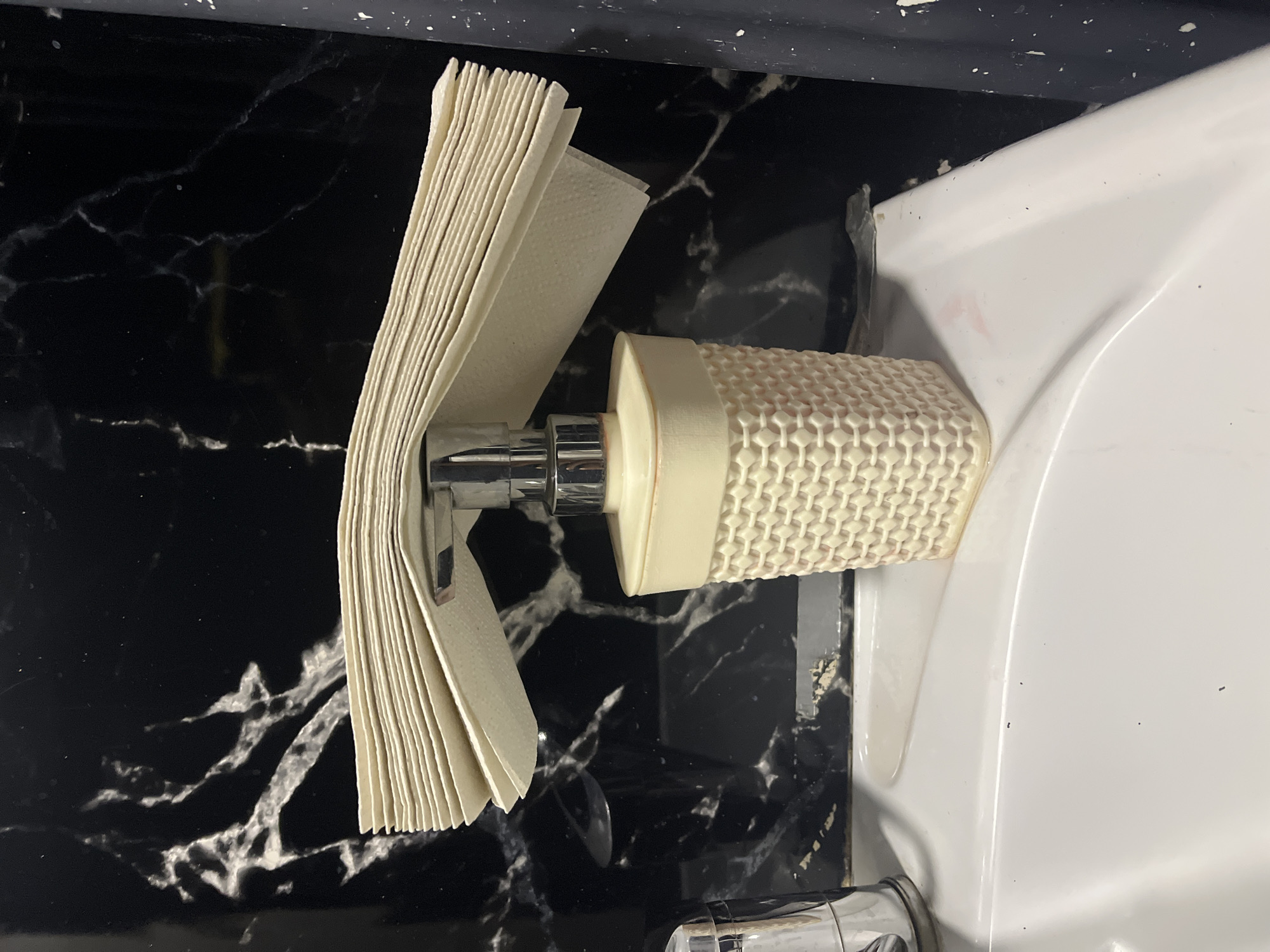 A close-up, top-down shot shows a stack of rectangular, light-beige paper towels on a chrome dispenser fixed to a marbled black countertop next to a white bathroom sink. The dispenser has a honeycomb-textured, cylindrical component. The sink's curvature is visible on the right side of the frame, along with a portion of its chrome faucet near the bottom. The setting appears to be a bathroom, possibly in a private residence or commercial establishment. The location is Chaikovsky, Russia.
