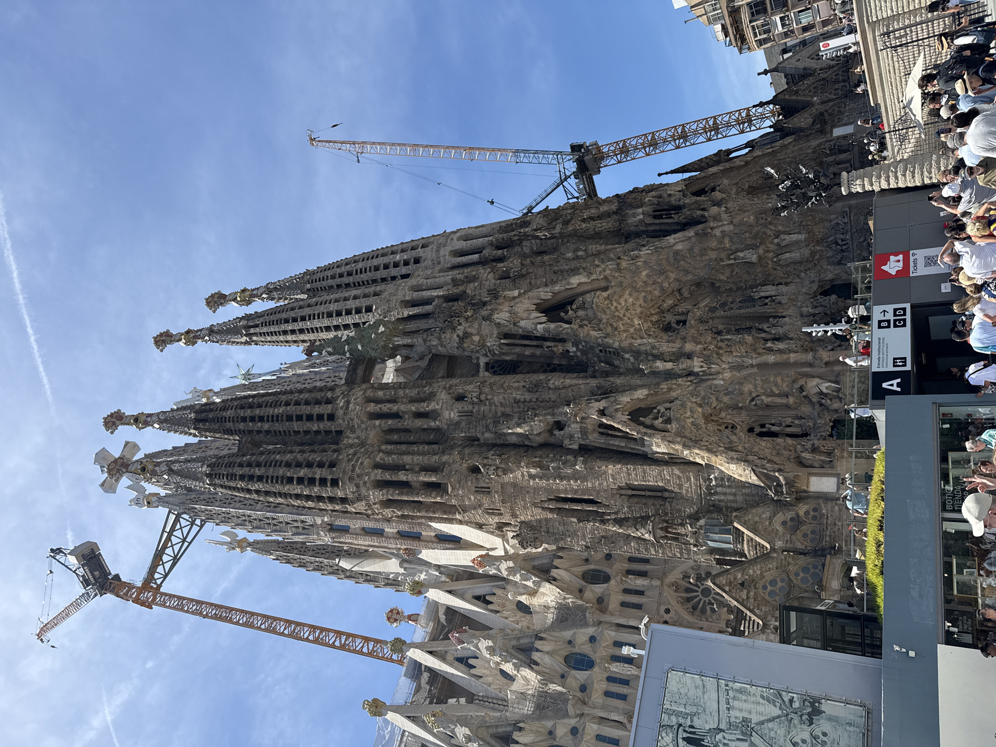 This image shows a low-angle, wide shot of the Sagrada Familia in Barcelona, Spain, on a sunny day with a blue sky and scattered clouds. Two tall construction cranes are prominently positioned next to the basilica, with one on the left and another on the right, indicating ongoing construction or restoration. The intricate, Gothic-inspired architecture of the basilica is visible, with its numerous spires and detailed stone carvings. Several people are seen gathered around the entrance of the basilica, with some walking and others standing. Signs with directions and ticketing information are also visible. The scene captures a blend of historical architecture and modern construction activity.