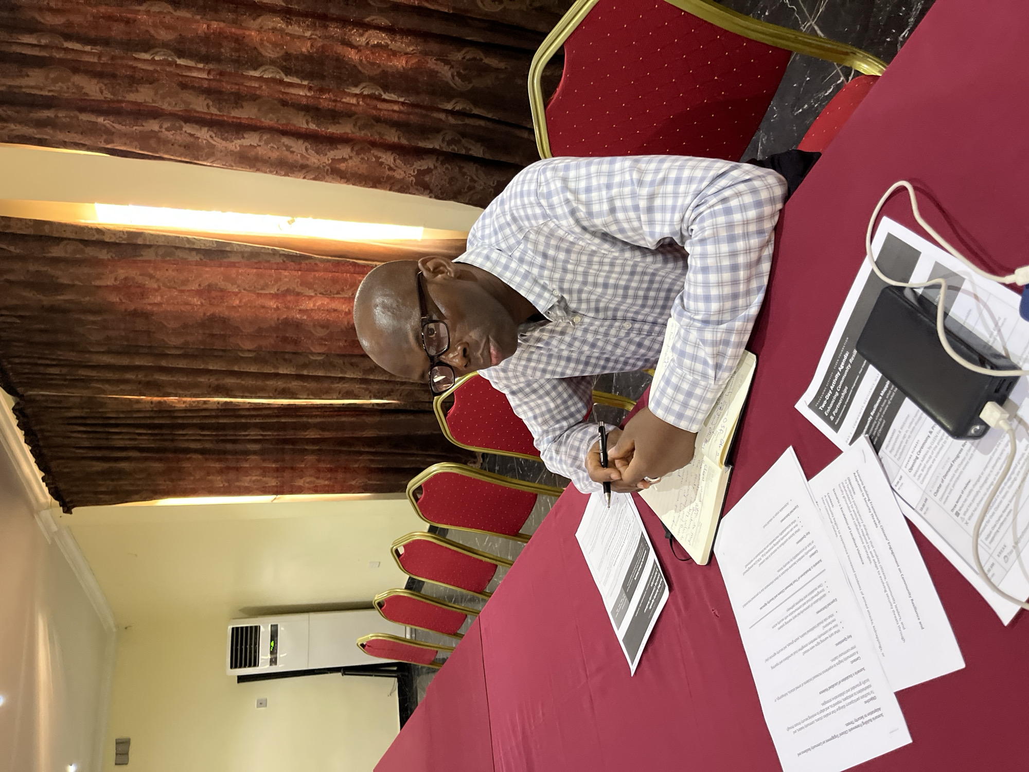 A man is sitting at a table in a room. He is writing in a notebook with a pen. There are papers, a smartphone and a charging cable on the table, alongside a number of red chairs and a red tablecloth. The room appears to be a meeting room with an air conditioner on the wall, and red curtains.