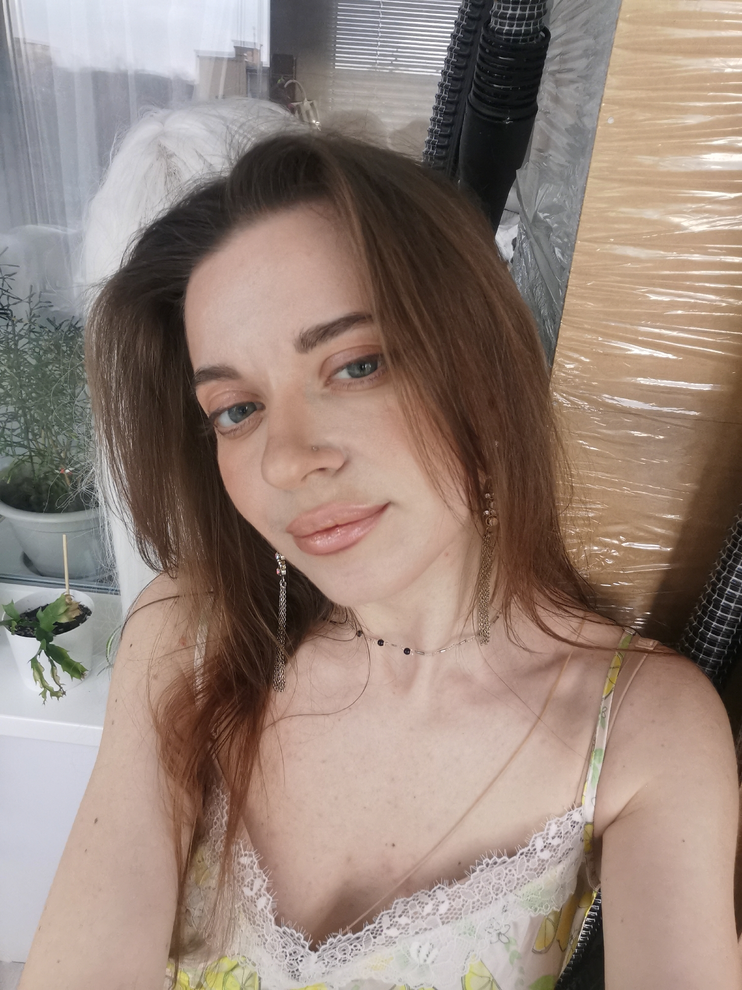 A medium close-up selfie shows a woman looking directly at the camera from an indoor setting. She has long brown hair, blue eyes, and a small stud nose piercing on her left nostril. Her lips are a glossy pink, and she wears a neutral expression. She is adorned with long, dangling chain earrings and a delicate necklace with small dark beads. Her attire appears to be a light-colored camisole or slip with white lace trim, possibly featuring a subtle yellow or floral pattern. Freckles are visible on her chest and shoulders.
The background to her left features a window with diffused natural light, suggesting daytime, possibly overcast. Two potted plants are visible on a windowsill: a larger, bushy green plant and a smaller succulent-like plant. The right side of the background is cluttered, showing a dark, corrugated tube or pipe, horizontal blinds, and a large, textured surface that resembles cardboard or wood wrapped in clear plastic film, creating a crinkled, reflective appearance. Below this, a checkered fabric or material is partially visible. No text is visible in the image.