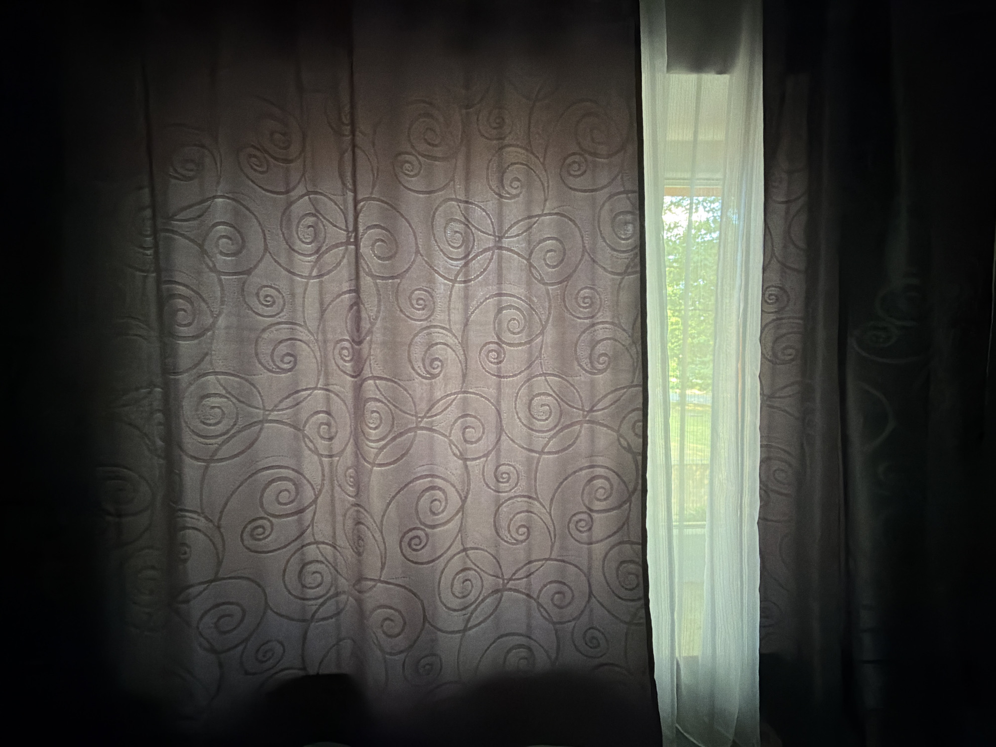 The image depicts a window covered by curtains in Dorsten, Germany. The window is partially obscured by two layers of curtains. The left curtain is a darker, textured fabric with a subtle swirling pattern. To the right of this, a sheer, white curtain is drawn back, revealing a view of the outdoors. Through the sheer curtain, greenery and trees are visible, suggesting it is daytime and likely fair weather. The lighting indicates that the sun is shining, casting a bright glow through the sheer fabric. The overall mood is somewhat subdued due to the contrast between the dark, heavy curtains and the bright light from the window, creating a sense of quiet and perhaps a hint of mystery. There is no visible text in the image.