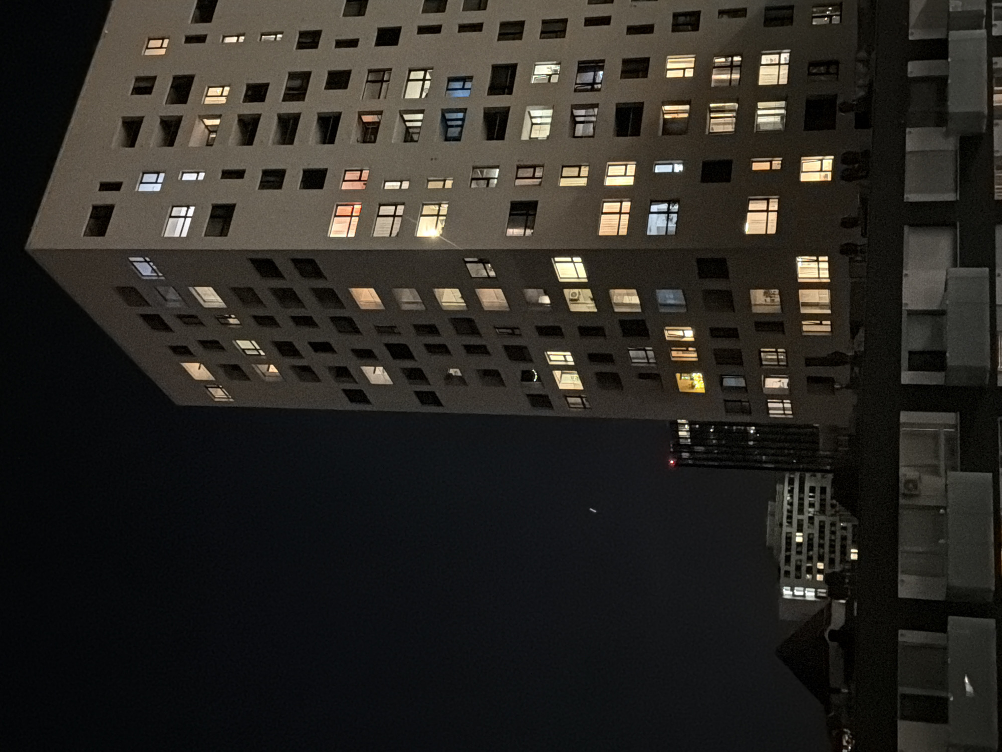 The image captures a nighttime scene featuring a tall, angular building with numerous windows illuminated from within. The building appears to be multi-storied, and its architecture is distinct, with a seemingly cantilevered section that overhangs the lower portion. To the right, there is another building partially visible, characterized by its modular design and vertical columns. The sky is dark and mostly clear, with a few scattered lights. The scene suggests an urban environment, possibly a city center or a densely populated residential area.