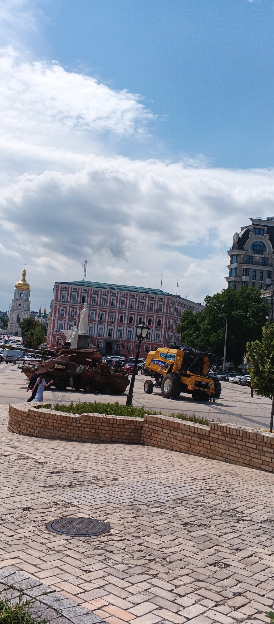 In Kyiv, Ukraine, on a sunny day with scattered clouds, the scene depicts an outdoor plaza paved with brick. In the foreground, a low brick wall borders a patch of grass. Further into the plaza, two distinct objects are on display: a damaged, rusted tank, and a yellow New Holland combine harvester. The tank appears to be a Russian T-64 or T-72 variant, showing signs of combat damage, including a missing turret and a torn hull. A woman in a light blue dress is captured in motion, seemingly taking a selfie with the damaged tank. Behind these objects, various buildings are visible, including a large red and white building with numerous windows and a golden-domed bell tower to its left, characteristic of Ukrainian Orthodox architecture. To the right, a modern building with a dark mansard roof stands tall. Cars are parked in the background, and a streetlamp stands between the tank and the combine harvester. The overall impression is a display of historical and agricultural machinery within a city square.