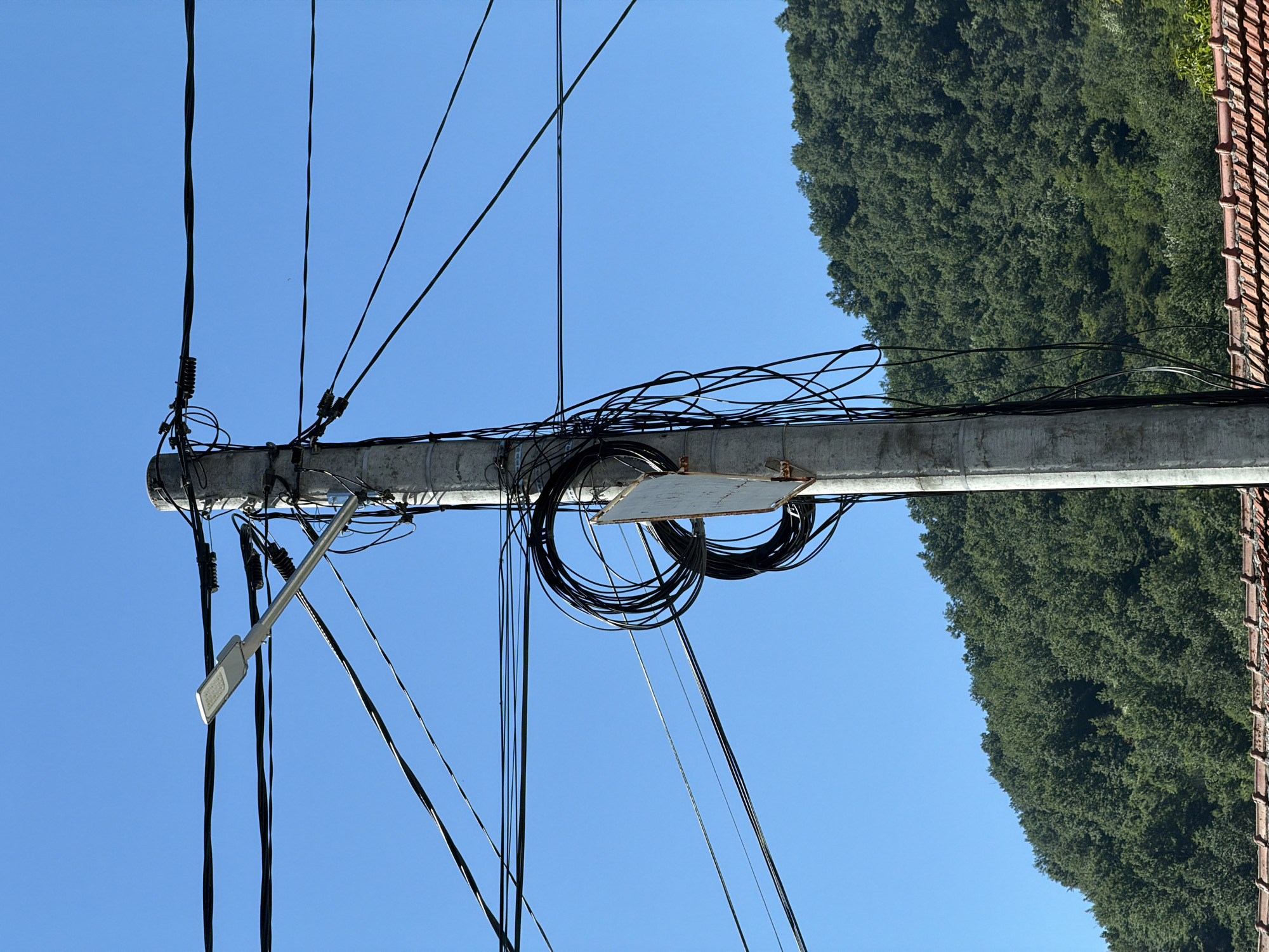 In the city of Beli Osam, Bulgaria, a concrete utility pole stands against a clear blue sky. The pole is adorned with a multitude of black electrical wires, some of which are neatly coiled, while others hang loosely. A modern street lamp is attached to the pole. Further down the pole, a white, rectangular sign is mounted. In the background, a dense, green forest covers a hillside, and the terracotta roof tiles of a building are visible at the bottom of the frame.