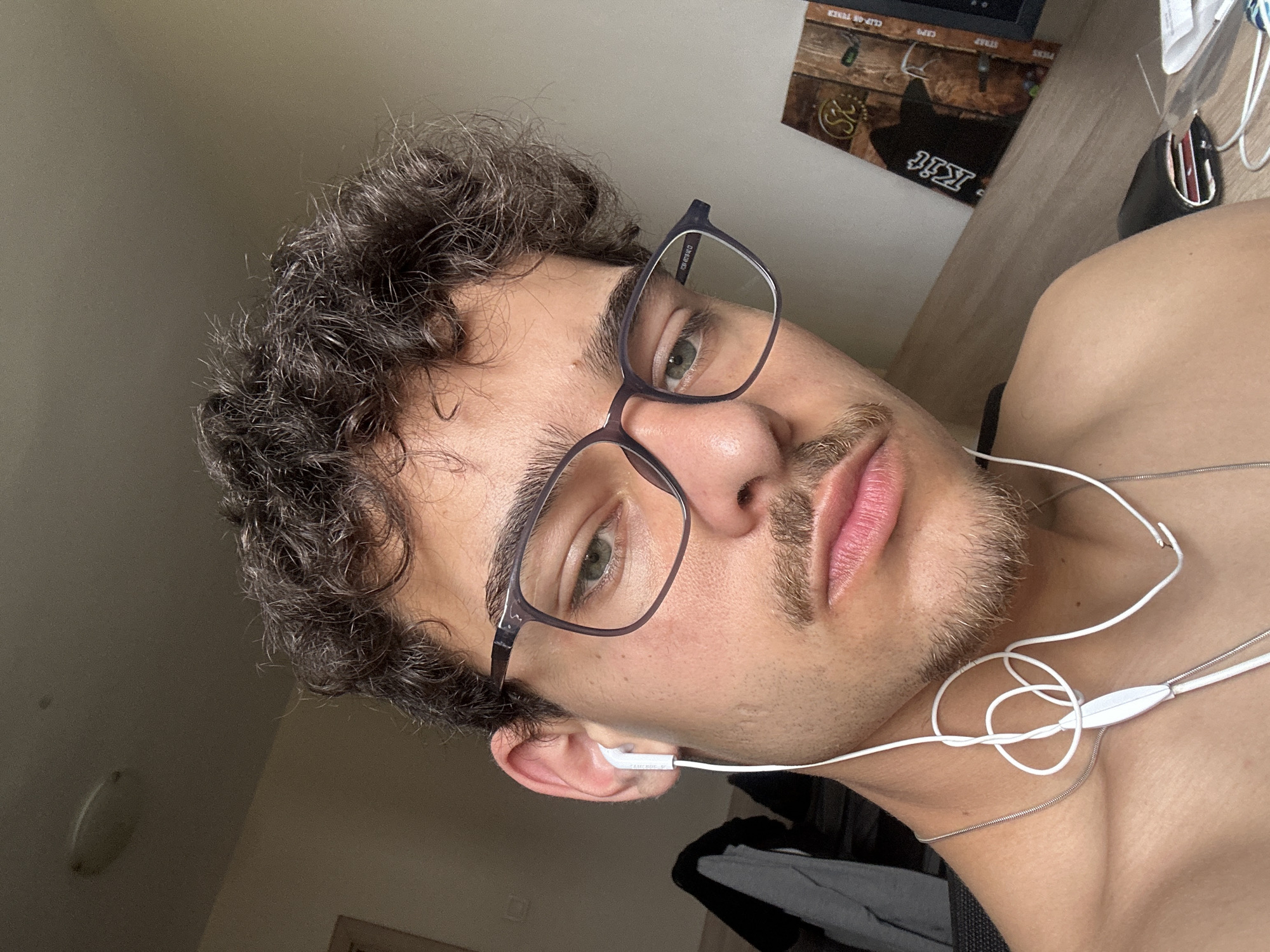 A young man with curly brown hair and a beard, wearing glasses and white wired earphones, is looking at the camera. He is inside, possibly a bedroom, given the desk with a keyboard, a guitar pick box, and headphones visible in the background. The location is not discernible from the image. There is no indication this image was taken in Sofia, Bulgaria.