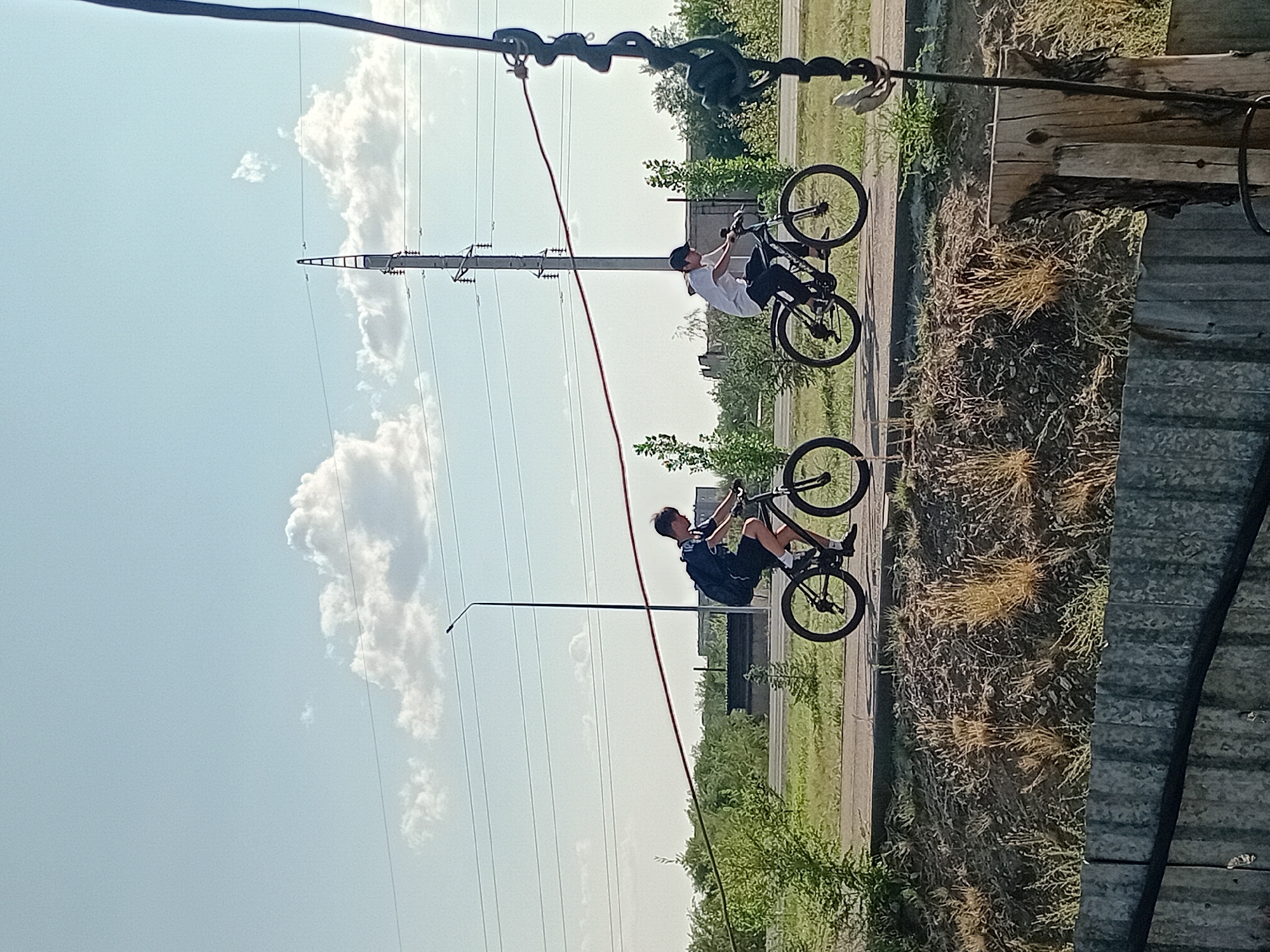 Two young males are cycling on a paved path. The male on the left wears a dark t-shirt with a white logo, dark shorts, and a dark backpack. The male on the right wears a white t-shirt, dark pants, and a dark baseball cap. They are cycling away from the camera, and the path runs parallel to a grassy embankment. In the background, there are trees, power lines suspended from utility poles, and a light blue sky with scattered white clouds. The scene appears to be on the outskirts of the city of Semey, Kazakhstan, possibly near a road or industrial area, given the presence of power infrastructure.
