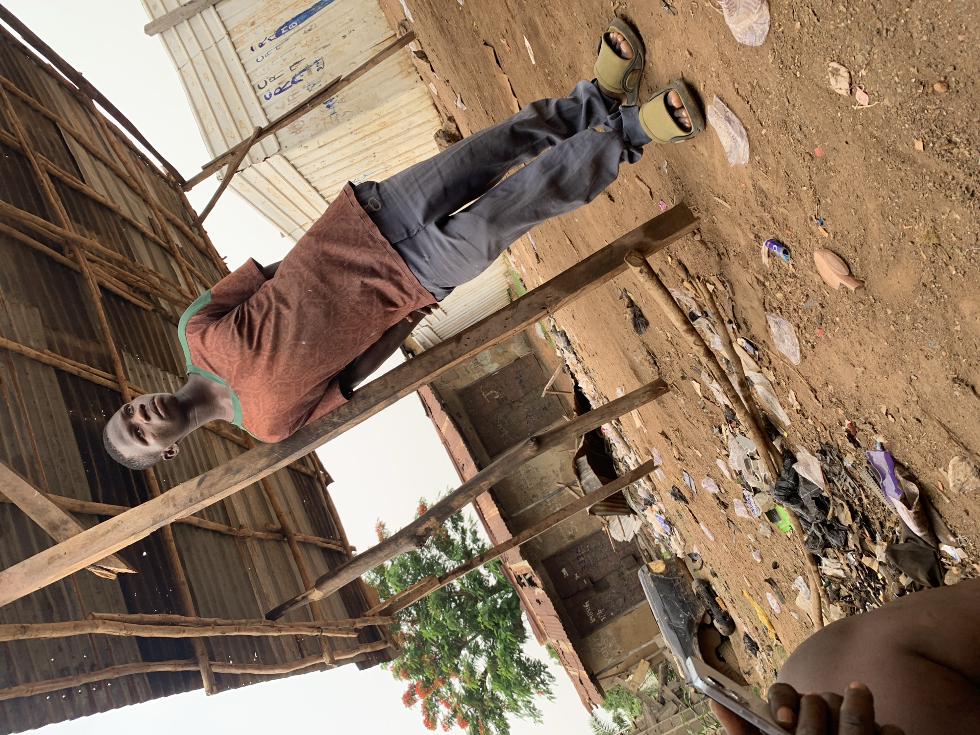 An eye-level outdoor shot captures a young man standing amidst makeshift structures in what appears to be a settlement in Akwanga, Nigeria. The man, positioned centrally, wears a brown patterned short-sleeved shirt with green trim, dark grey trousers, and light green open-toed sandals. He looks directly at the camera with a neutral expression.
In the immediate foreground, to the bottom right, part of another person's dark-skinned arm and hand holding a silver smartphone is visible, suggesting the photo was taken from a lower perspective.
The setting is characterized by unpaved, reddish-brown dirt ground, strewn with various forms of litter including plastic bags, discarded sandals, and a distinctive wooden fish-shaped object. The area features simple, ad-hoc constructions: corrugated metal sheets are used for roofs and walls, supported by rough wooden beams and poles. One white corrugated ceiling section, visible in the upper right, has blue writing on it, which includes characters resembling "بسم الله" (Bismillah - In the name of God) in Arabic script, along with other indistinct marks. Another dark corrugated structure in the background also shows some faint white writing, possibly "KOFE...".
Beyond the structures, a lush green tree with small red flowers is visible, contrasting with the man-made environment. The sky appears overcast and bright, indicating it is daytime. The overall scene suggests a glimpse into daily life in an area with basic infrastructure.