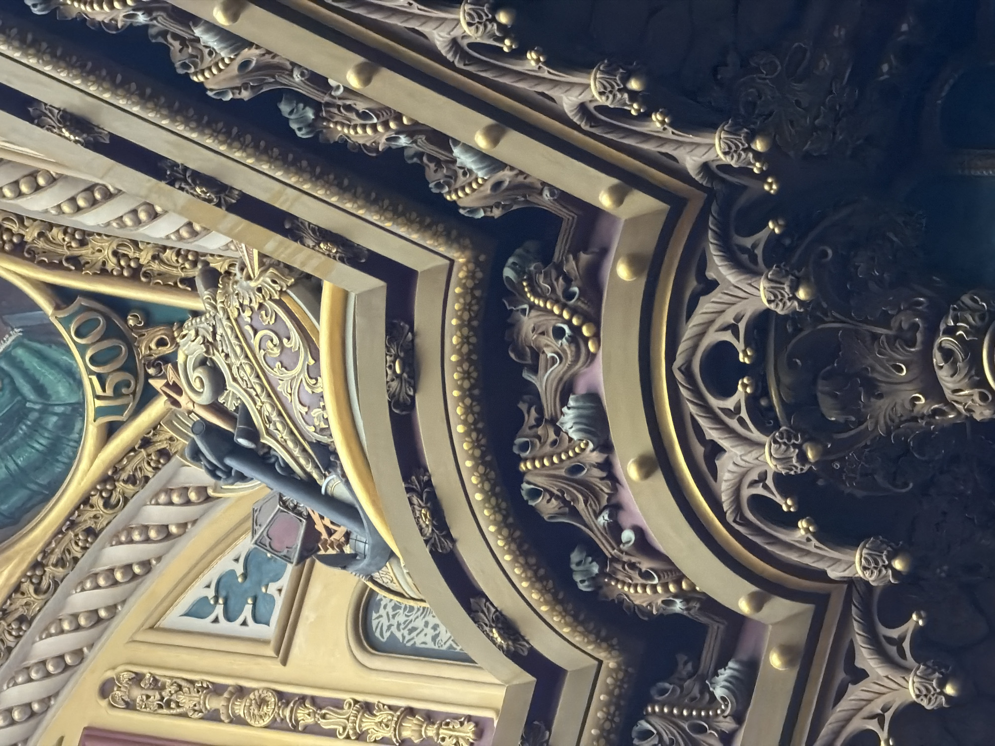 The image depicts a close-up view of ornate architectural detailing, likely part of a ceiling or upper wall, in a building that appears to be a historic or grand public space. The style is baroque or rococo, characterized by intricate carvings, gold embellishments, and rich colors, including deep purples, golds, and teal. Prominent features include detailed floral motifs, beaded garlands, and what appears to be an anchor and part of a ship's wheel, suggesting a maritime theme. The numeral "1500" is visible, possibly indicating a year or date significant to the building's history. The lighting is somewhat dim, highlighting the textures and three-dimensional qualities of the ornamentation. There are no people or animals visible in the frame.
This specific architectural detail is located in the historic center of Rio de Janeiro, Brazil, within the Candelária Church. The Candelária Church, a prominent landmark, is known for its impressive Baroque and Neoclassical interior. The ornamentation visible in the image is consistent with the detailed and opulent style found within the church's sanctuary and dome.