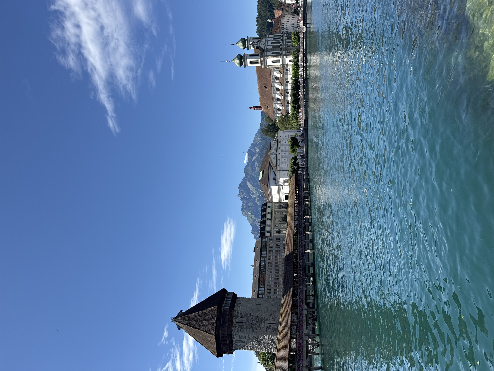 The image shows a view of the Reuss River in Lucerne, Switzerland, on a clear, sunny day. In the foreground, the turquoise water of the river reflects the blue sky and surrounding architecture.
Dominating the left side of the image is the Kapellbrücke (Chapel Bridge), a historic wooden covered bridge that spans the Reuss. The bridge features an octagonal water tower, Wasserturm, made of stone, with a conical wooden roof. The bridge itself is a long, covered wooden structure supported by piers, with what appear to be flower boxes along its sides.
To the right of the bridge, a row of buildings lines the riverbank. Most notable among them is the Hofkirche (Court Church), a grand white Baroque church with two green-domed towers and a red-tiled roof. Adjacent to the church is a larger, classical-style building with a white facade and a visible red roof. Further in the background, mountains are visible under the clear blue sky, with a few wispy clouds.
Several people are visible walking along the riverbank and on the bridge, suggesting a public area and tourist attraction. Birds are also visible in the sky. The overall scene depicts a picturesque and vibrant urban landscape with historical landmarks and natural beauty.