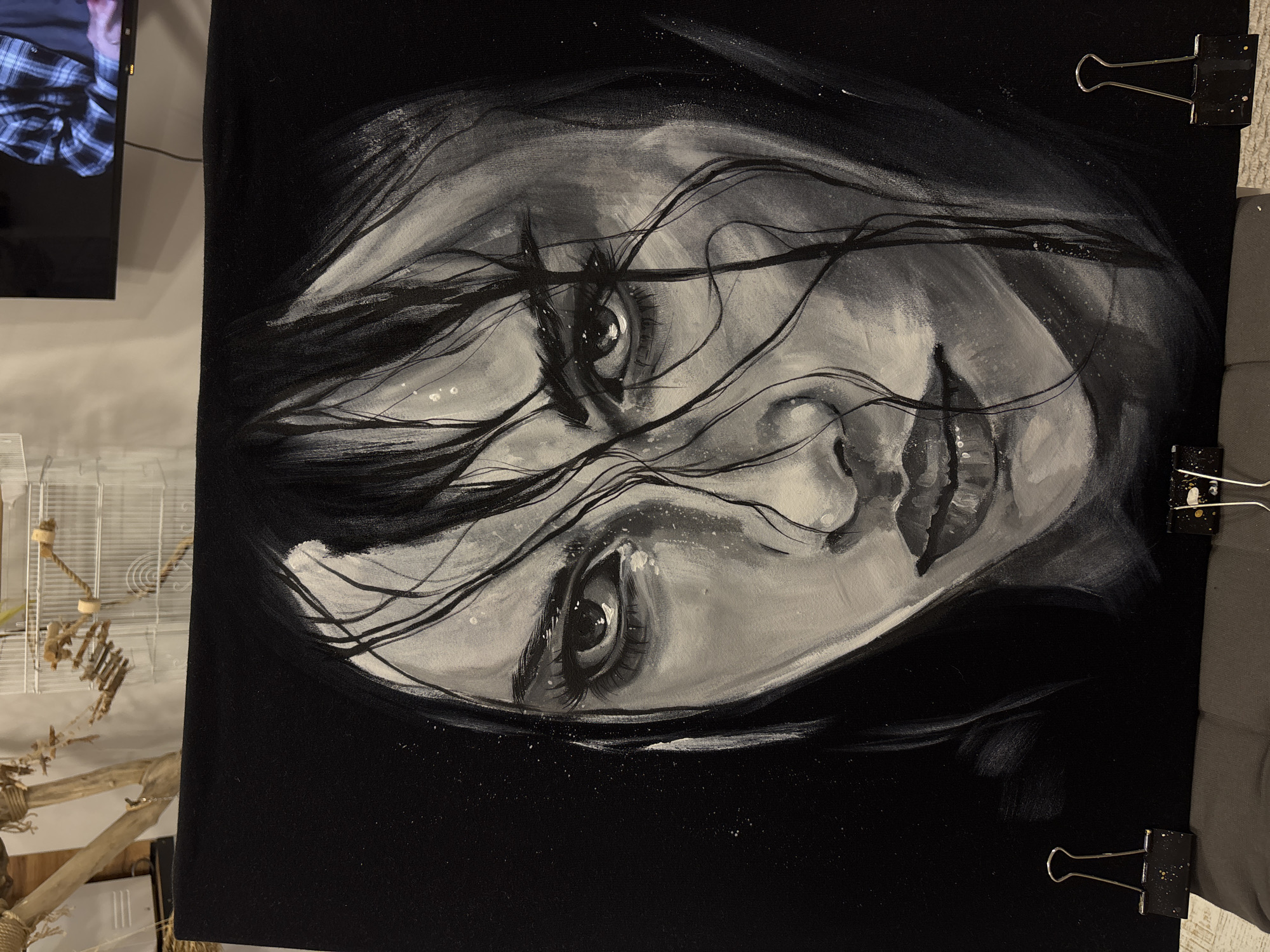 The image shows a monochrome painting of a woman's face, tilted sideways. The painting is mostly black and white, with visible brushstrokes. The woman has long hair obscuring part of her face. Her eyes are prominent and appear to be the focal point of the painting. The painting is clipped to something on the right side by two black binder clips. In the background, partially visible, is a bird cage, a TV showing a person in a plaid shirt, and what appears to be a beige-colored wall or floor. The location cannot be determined from the image alone and no information about Krasnoyarsk, Russia is present.